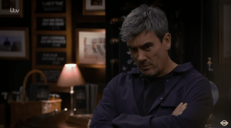 Cain Dingle sits in the Woolpack with his arms folded looking brooding as Lydia investigates who killed Craig in Emmerdale