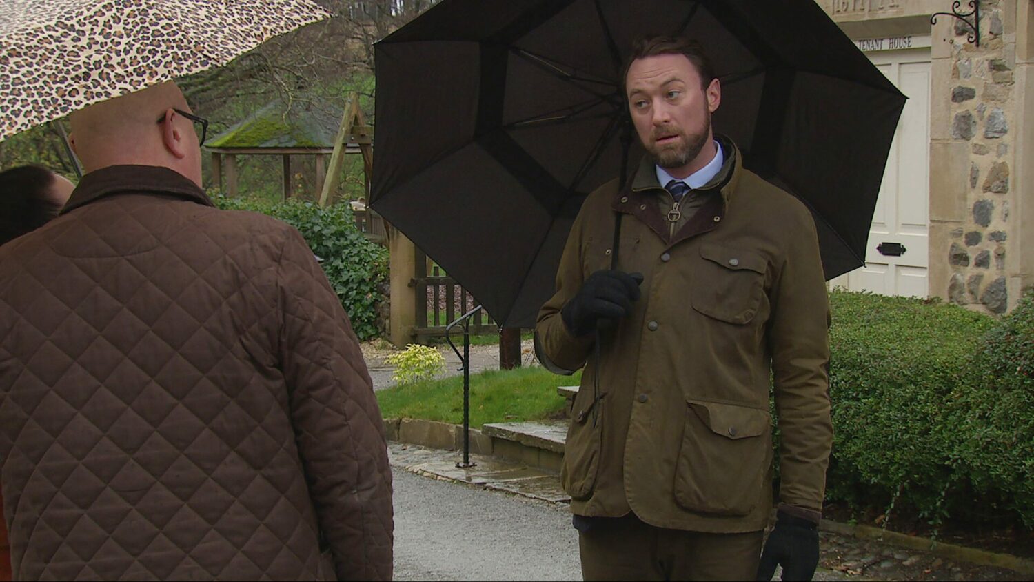 Liam in Emmerdale looks serious as he talks to Mandy and Paddy with umbrellas up.