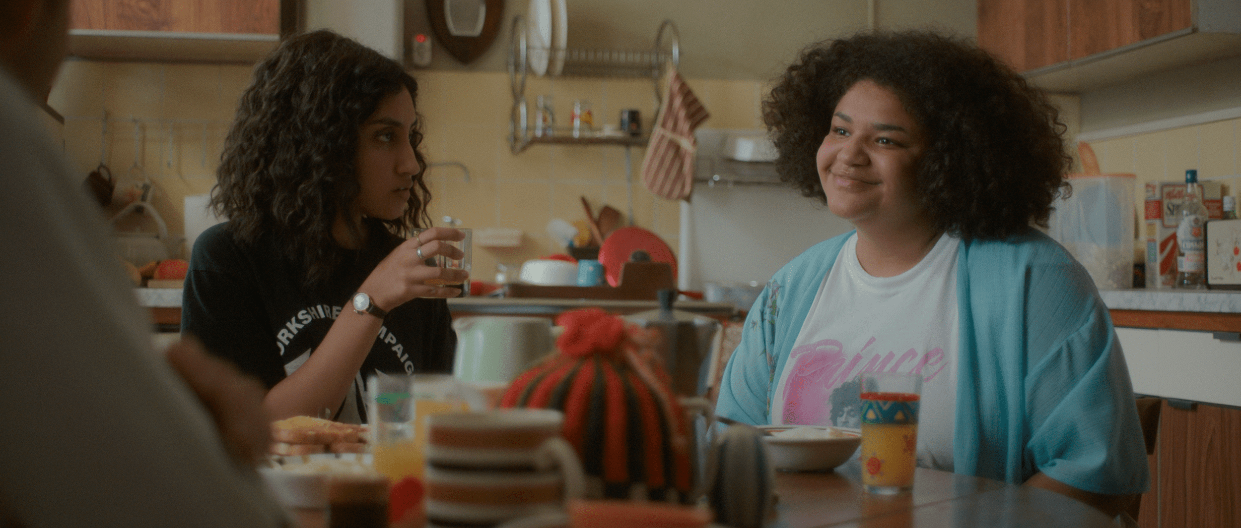 A smiling Tilly (Amber Grappy) at the dining table with Emma (Ambika Mod) in One Day