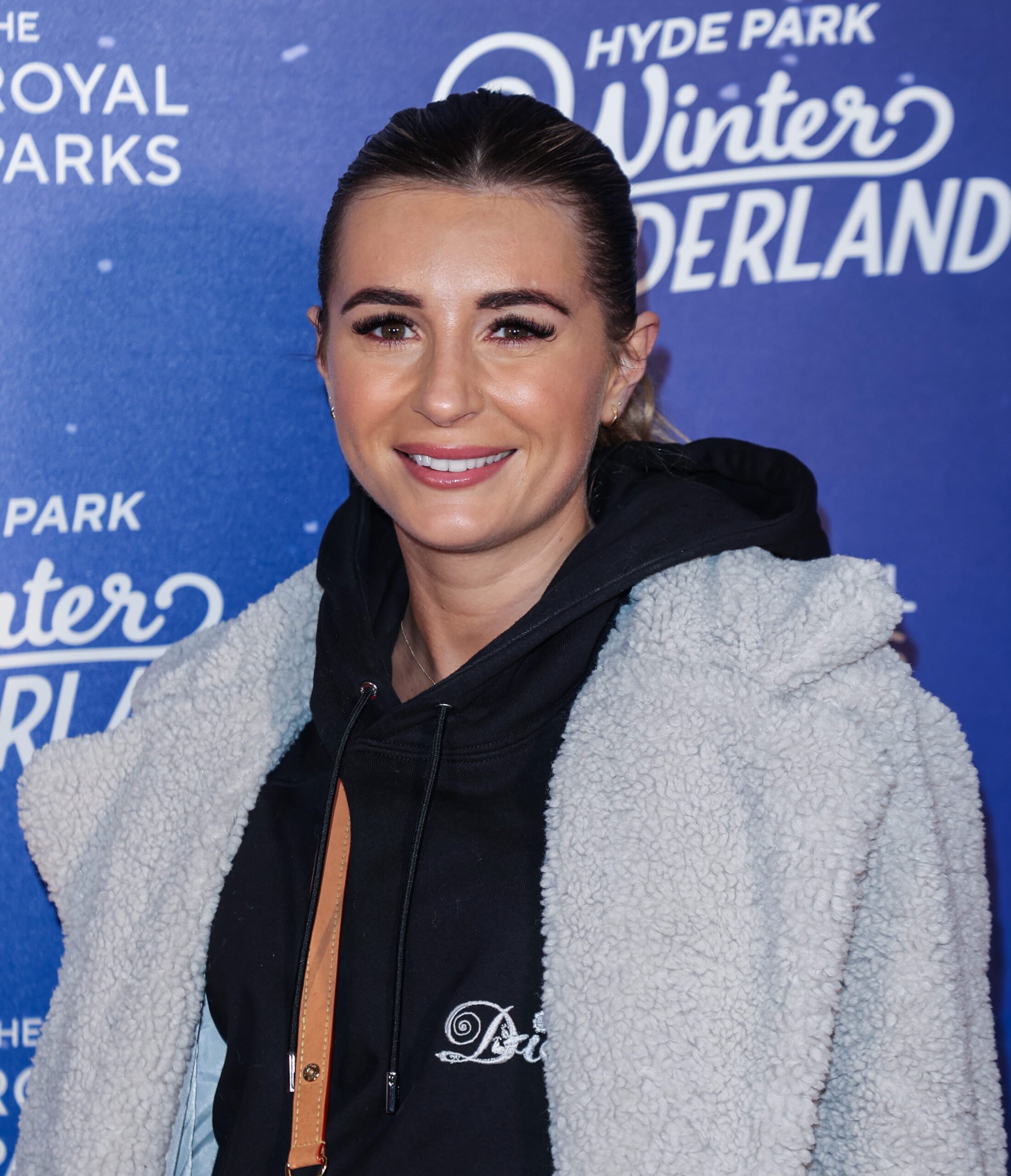 Dani Dyer smiling at the camera wearing her hair in a slicked back ponytail, a black hoodie and a grey coat