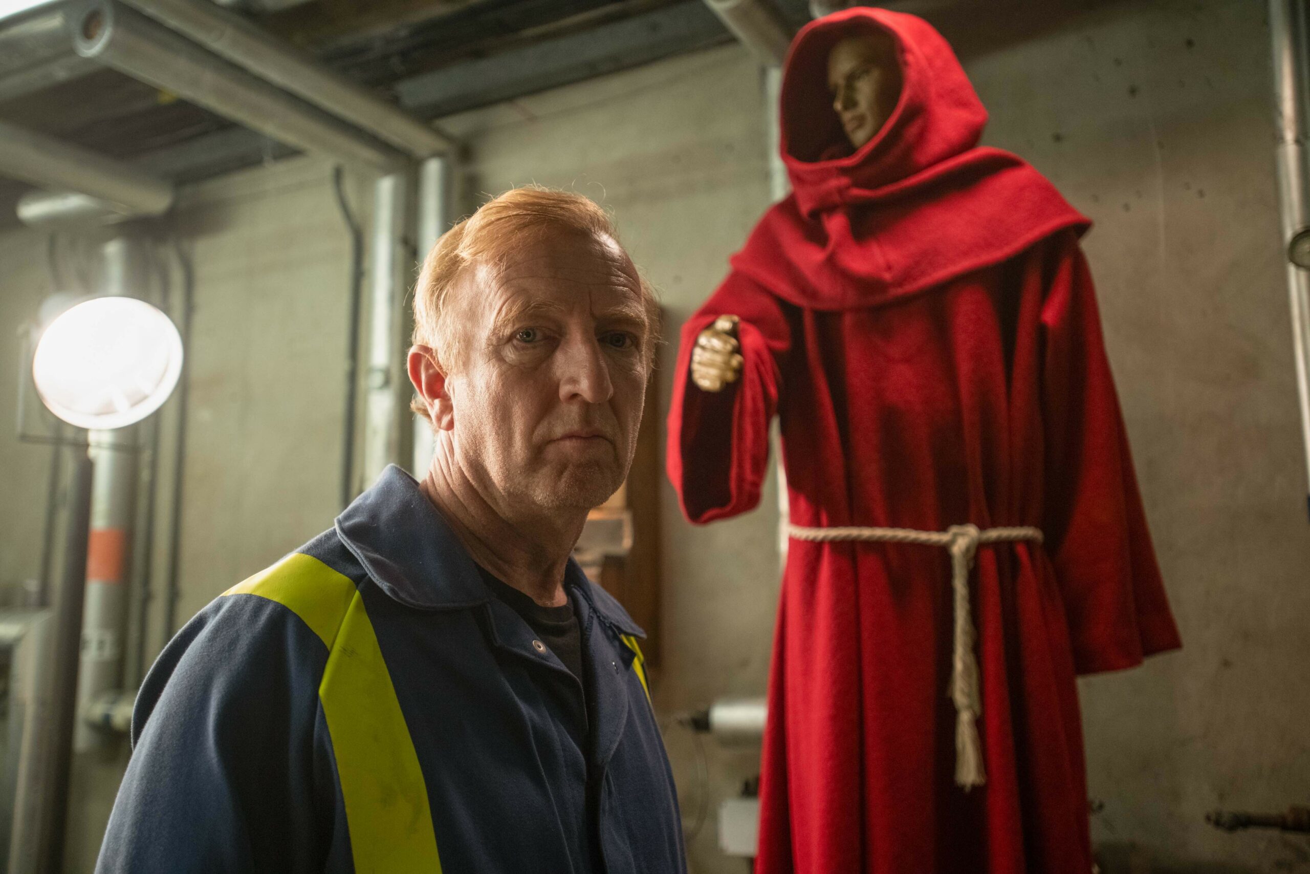 Steffan Rhodri as Geoff in The Way, wearing high viz, in front of a model of a monk in red robes