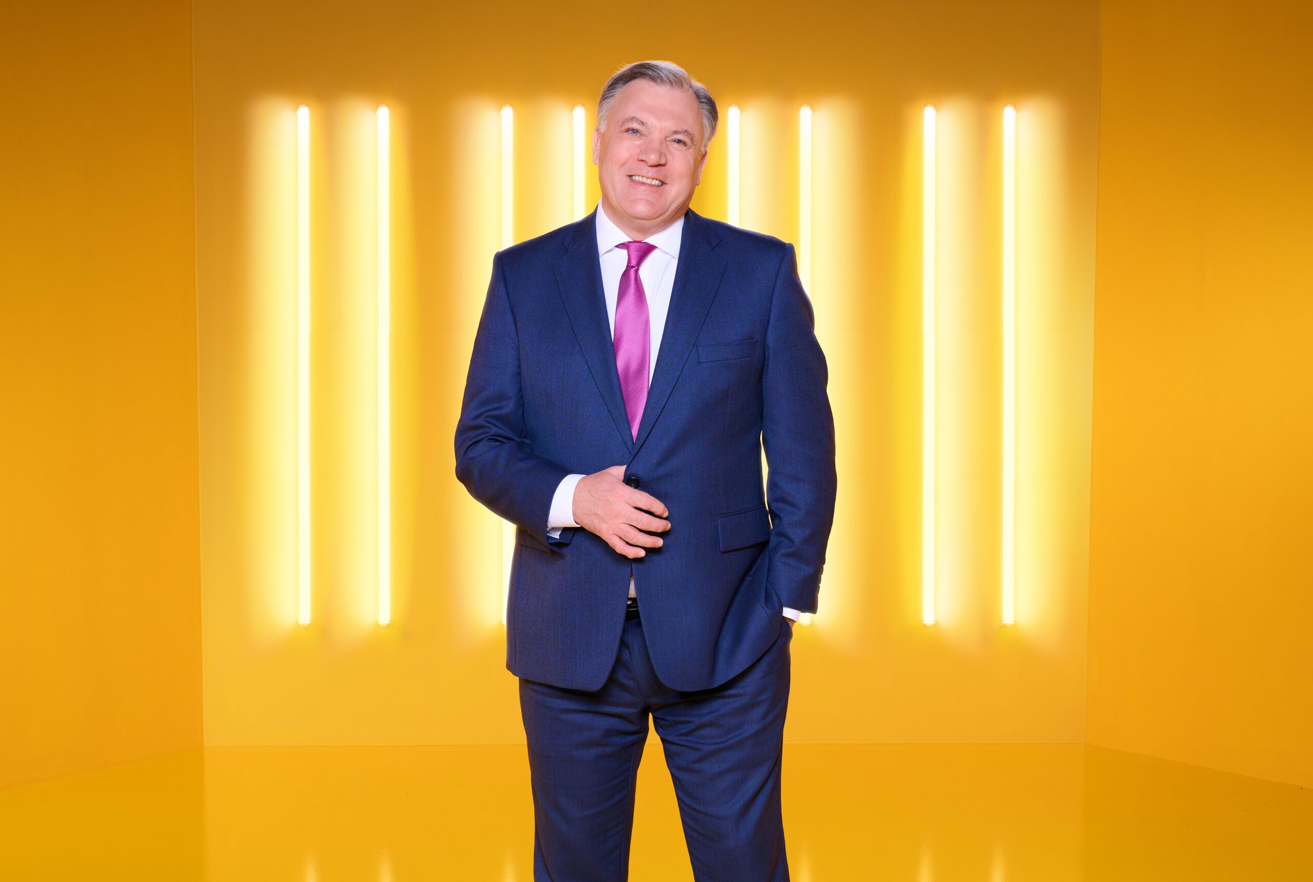 Possible Good Morning Britain co-host Ed Balls smiling while wearing a blue suit and pink tie, on a yellow background