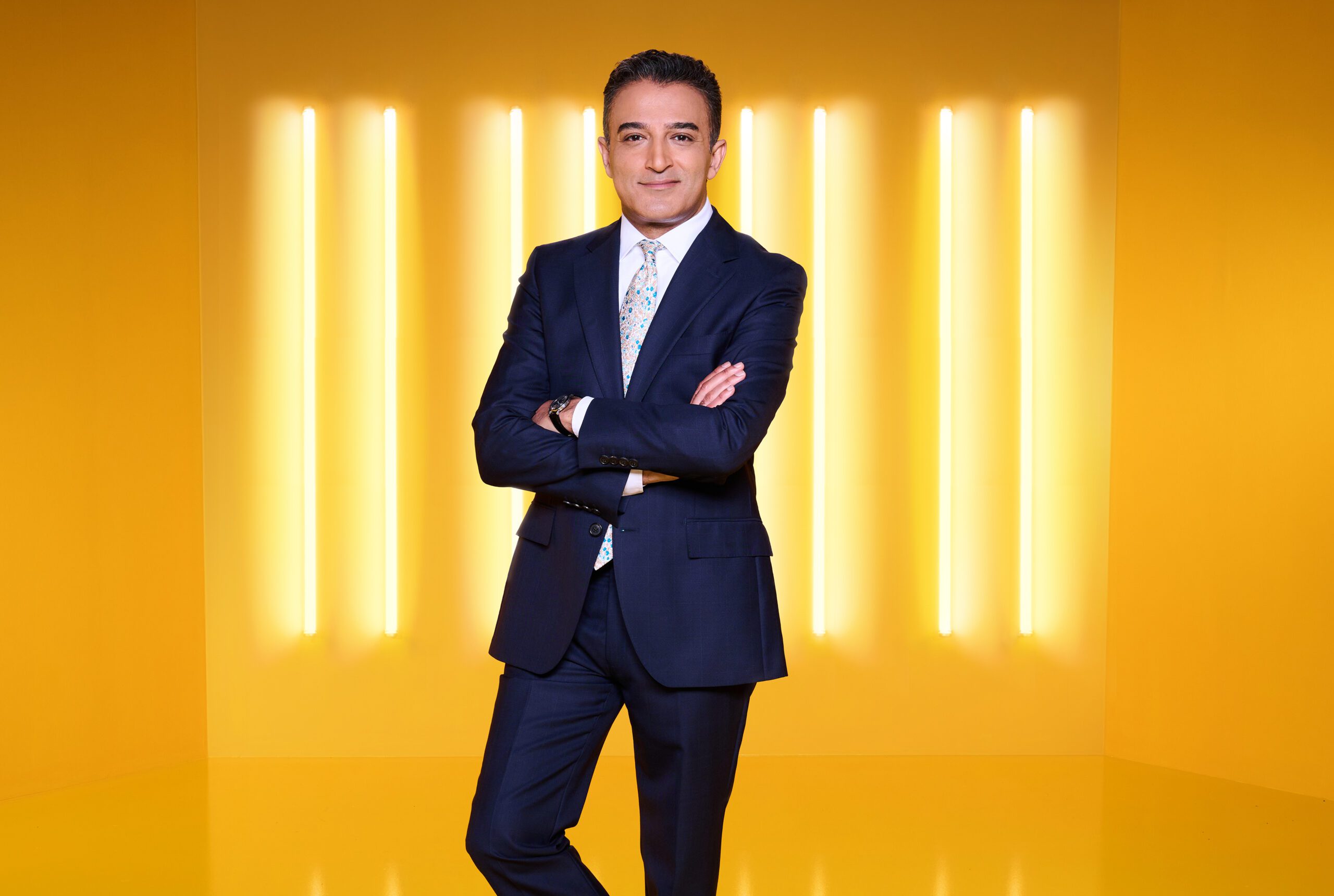Adil Ray smiling at the camera, wearing.a suit with light-coloured tie, arms crossed and leg bent. There is a yellow background.