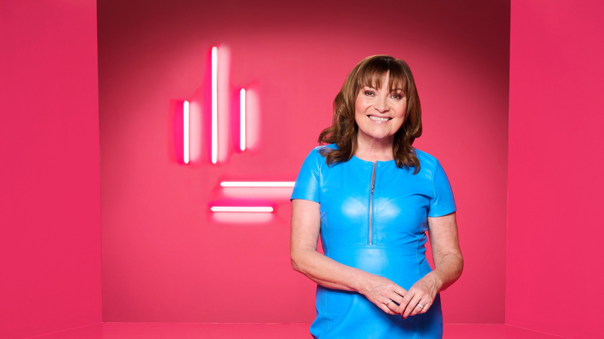 Lorraine Kelly wearing a blue dress standing in front of a pink background