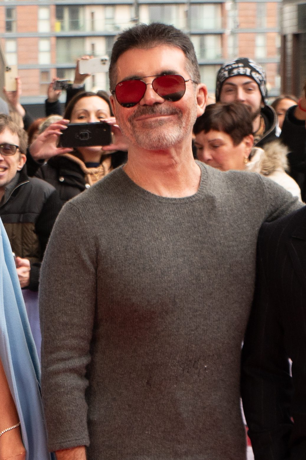 Simon Cowell arrives ahead of the 2024 Britain's Got Talent Auditions at the Lowry in Manchester in 2-24. He is wearing sunglasses and a grey shirt, and surrounded by fans.