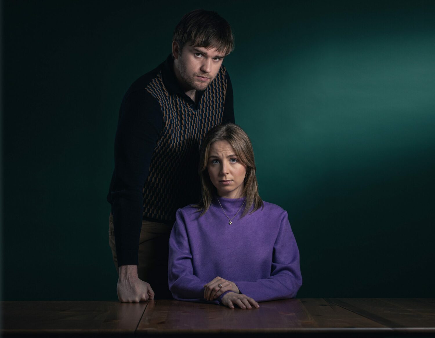 Promo shot of Tom King and Belle Dingle he's looming behind her looking aggressive while she sits at the table looking afraid