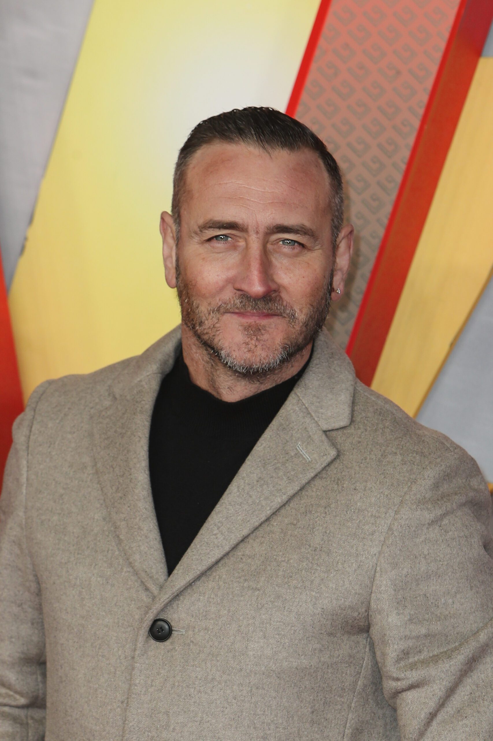 Will Mellor smiling on the red carpet in a beige jacket