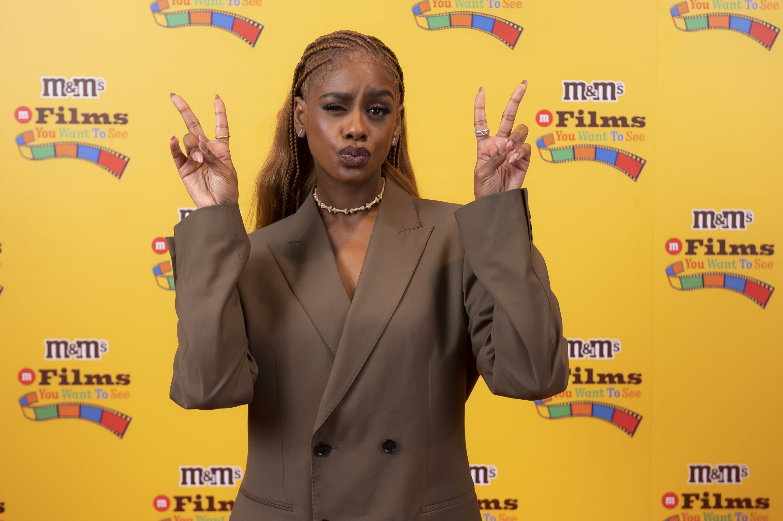 Zeze Millz wearing a brown blazer, making two peace signs with her hands, at the #FilmsYouWantToSee, created by M&Ms. 