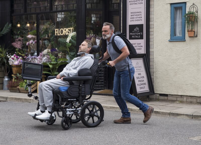 Billy pushes Paul's wheelchair through the street on Corrie