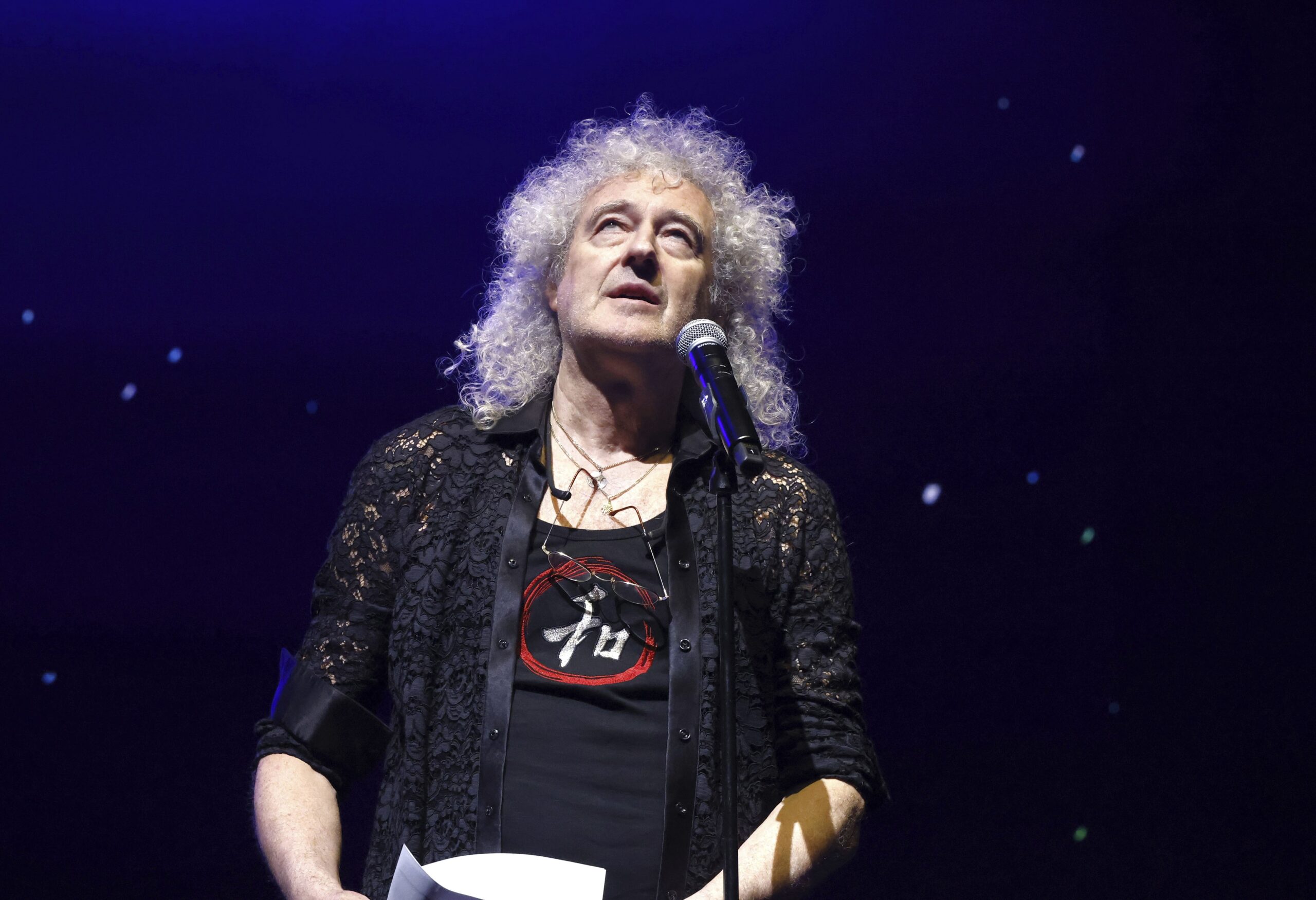 Brian May looking upwards in front of microphone on stage