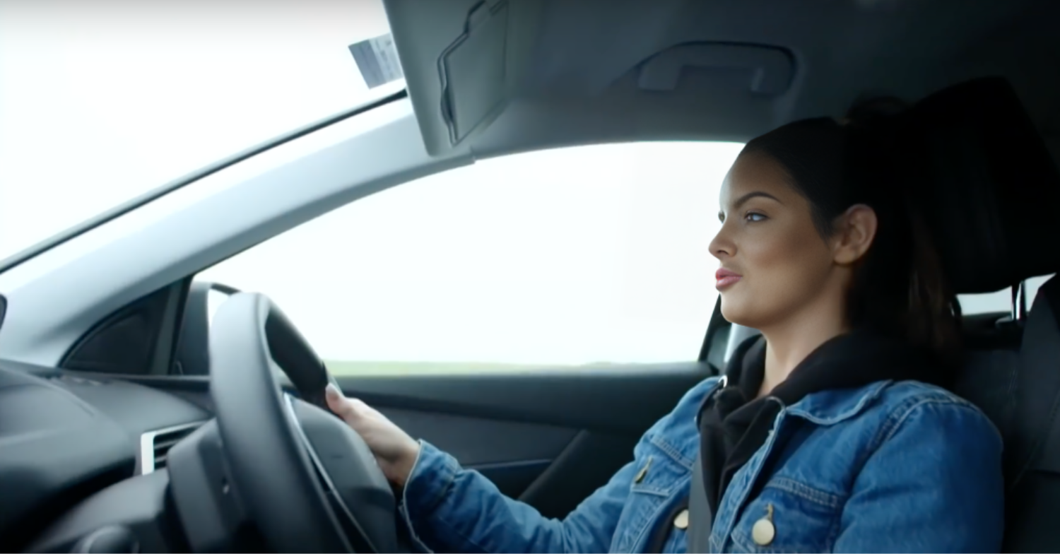 Maura Higgins purses her lips while driving a car in Maura's Ireland. She is wearing a denim jacket on holding the wheel.