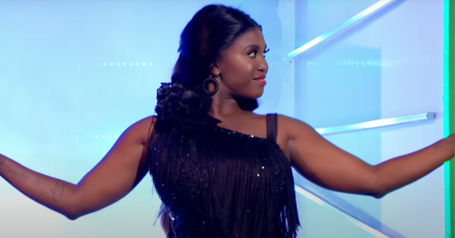 Motsi Mabuse has long black hair and is wearing a black dress. She has her arms up looking to the right while performing on Strictly Come Dancing.