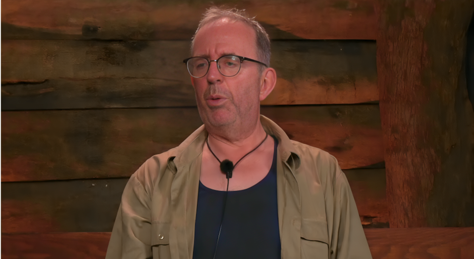 Reverend Richard Coles wears a black tee with a tan shirt, he is talking about GK Barry in the I'm A Celebrity confessional.