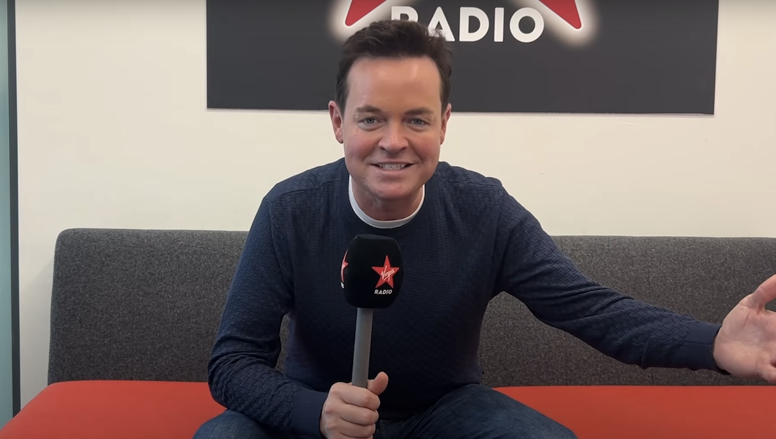 Stephen Mulhern holding a Virgin Radio mic to his mouth while sitting on a red sofa and gesticulating
