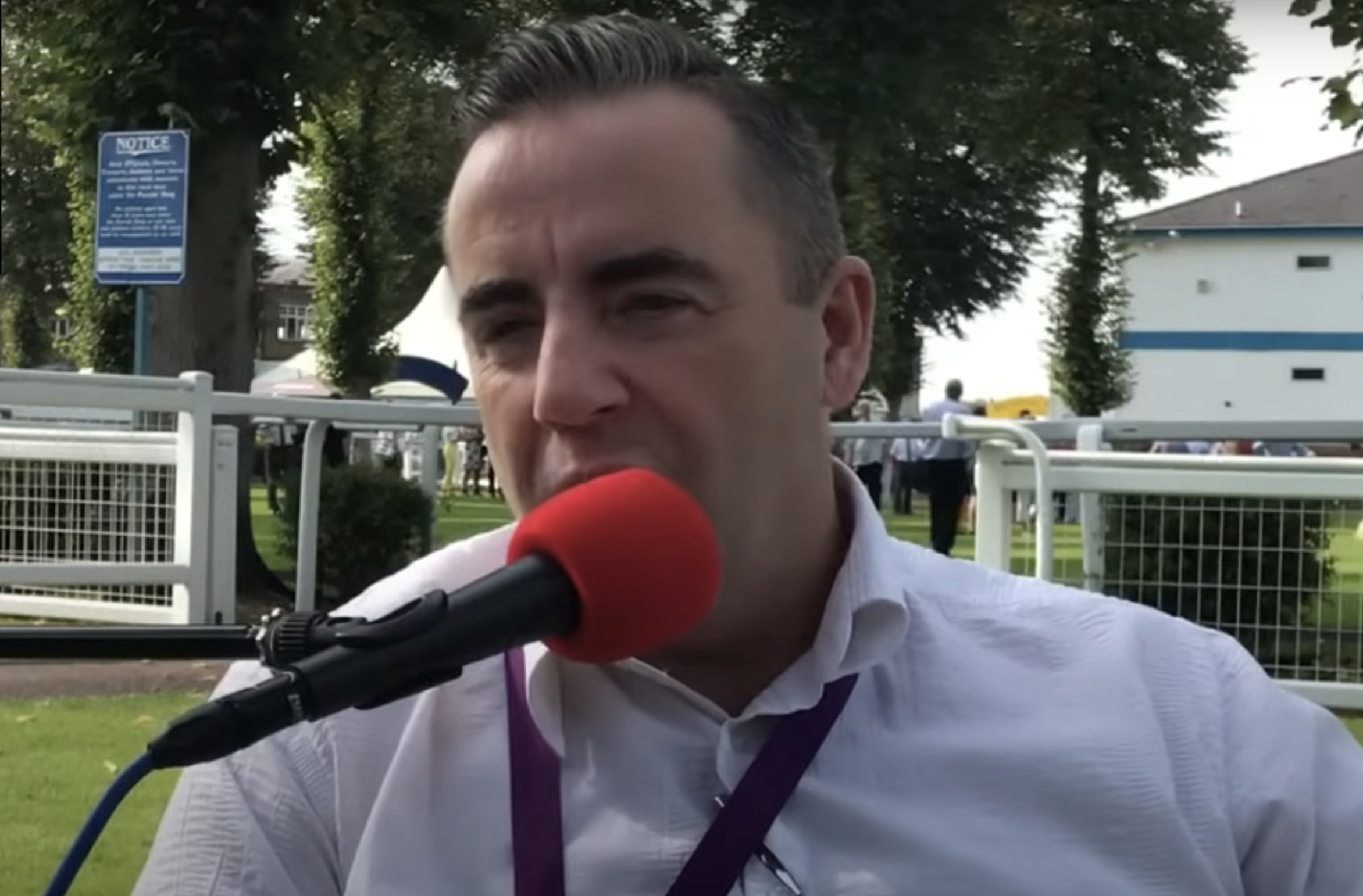 John Hunt in a shirt speaking into a microphone with a red cover