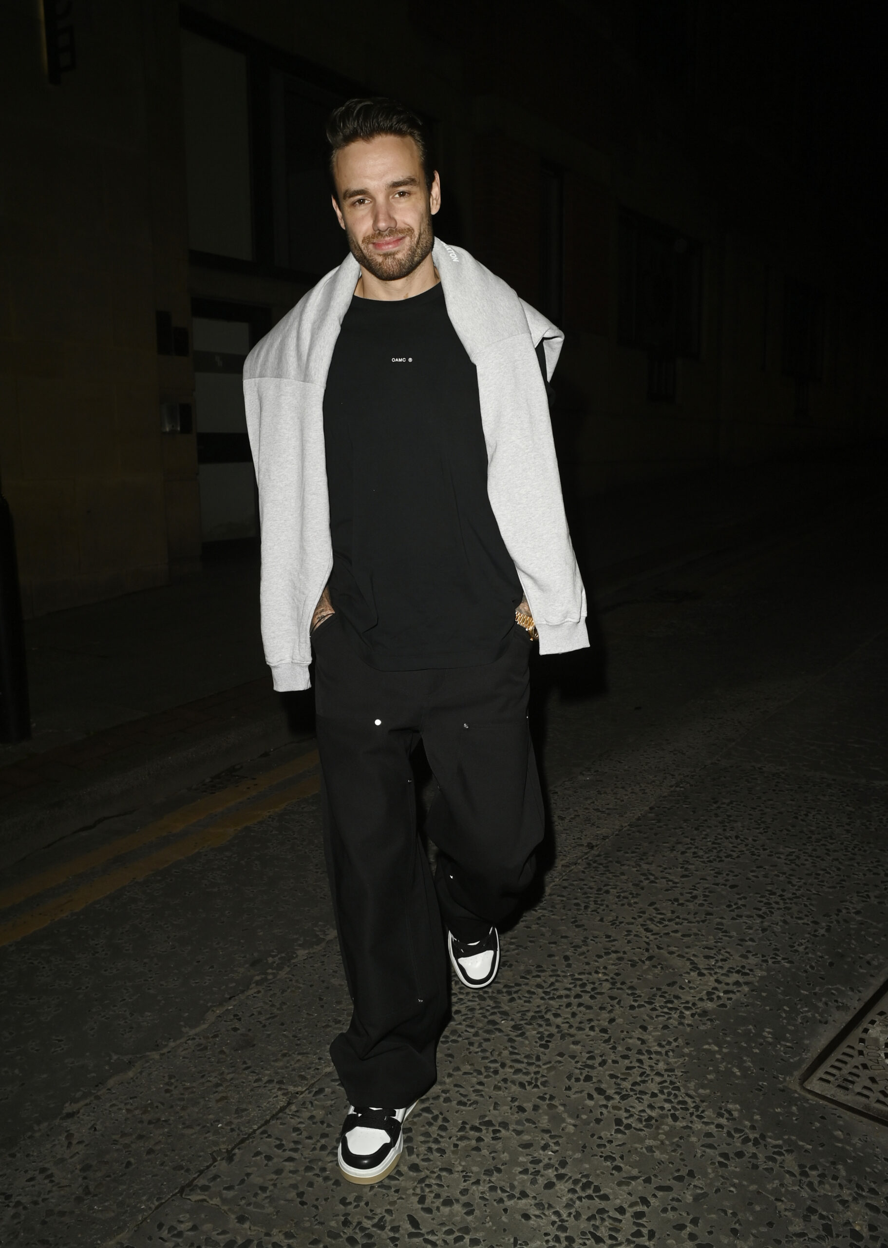 Liam Payne wears a black tee and sweatpants with a white jumper around his shoulders