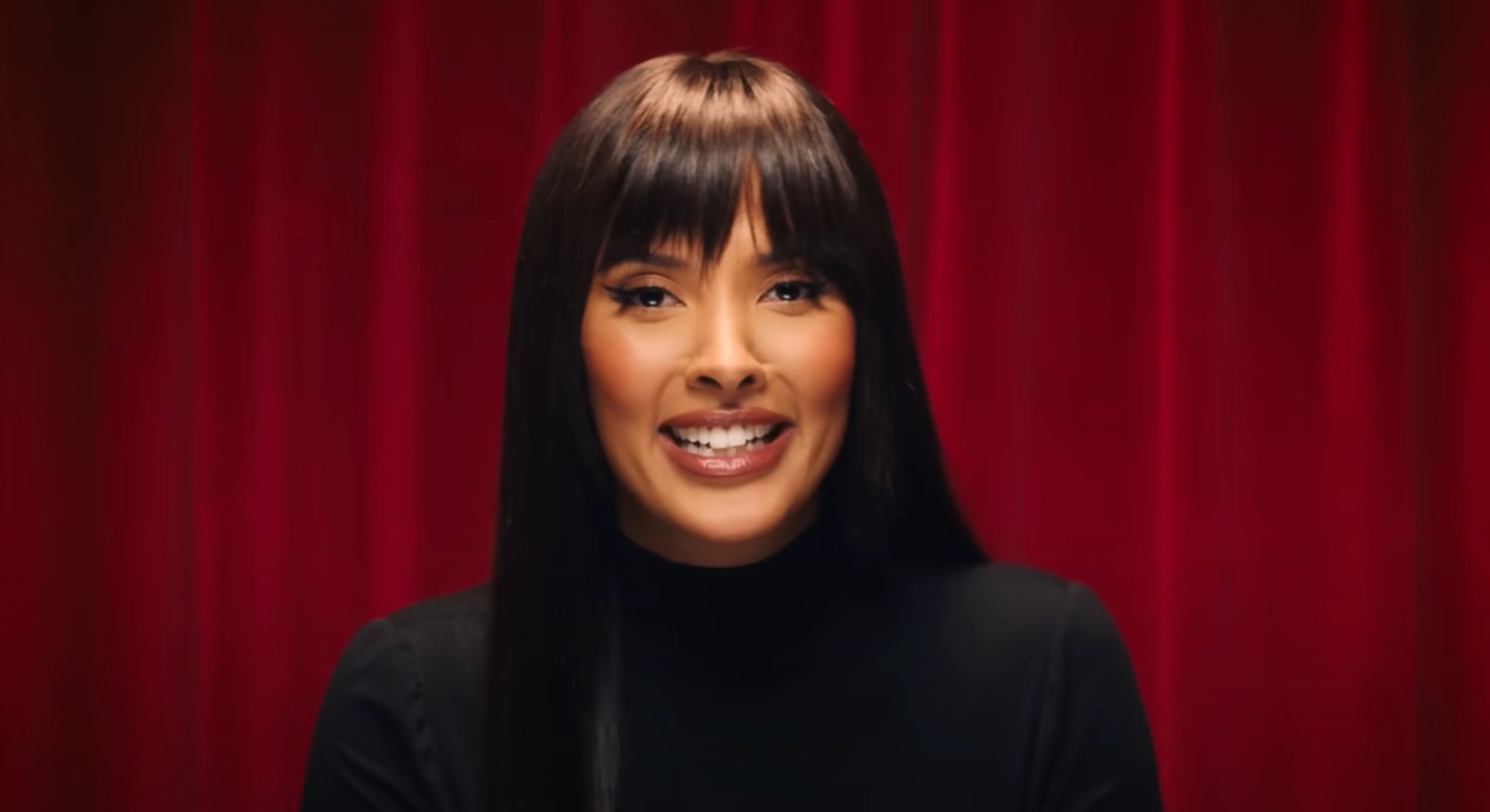 Maya Jama wears a black turtle neck top and sits in front of a red velvet curtain