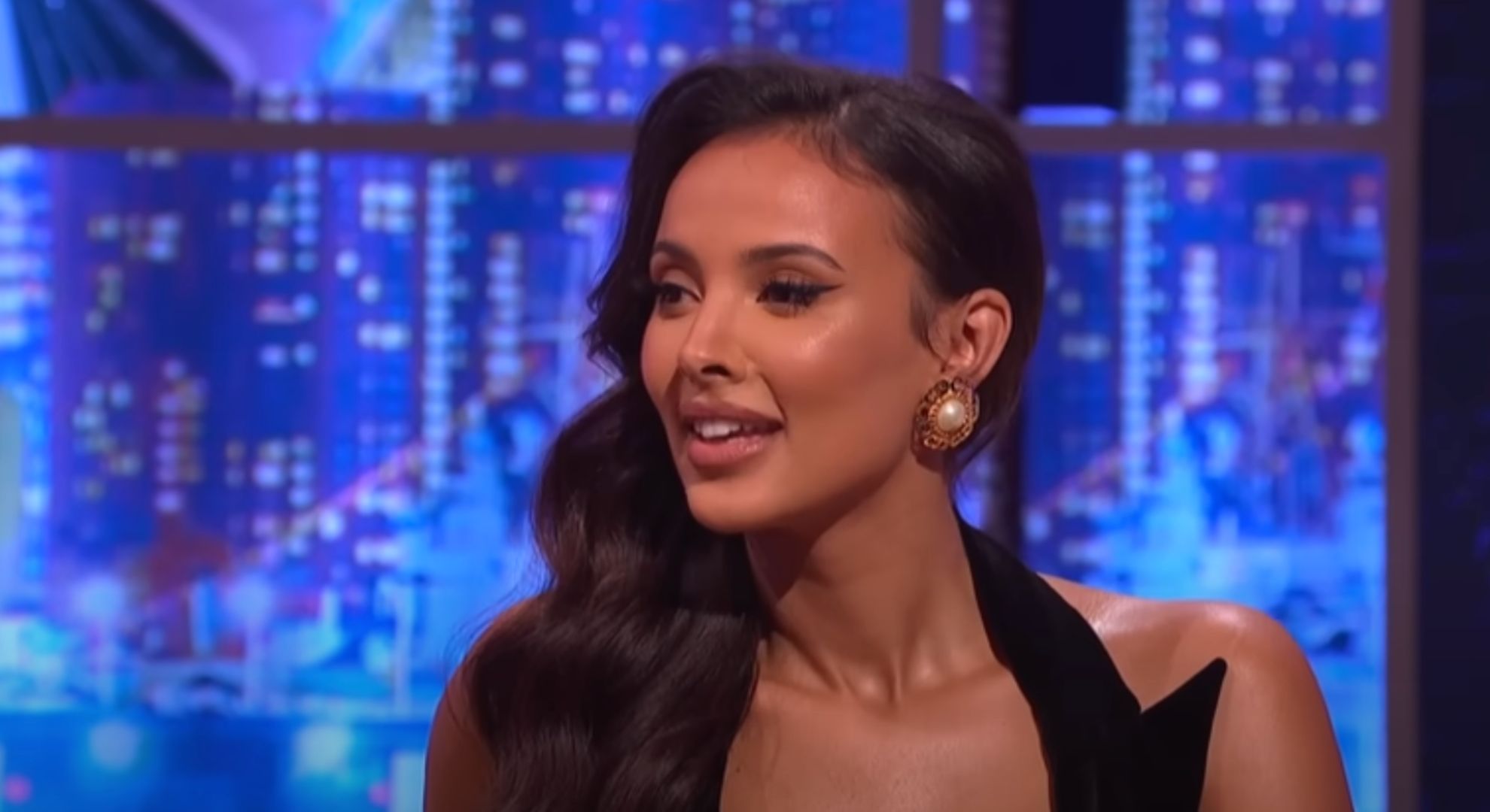 Maya Jama wears a black dress and sits on a sofa