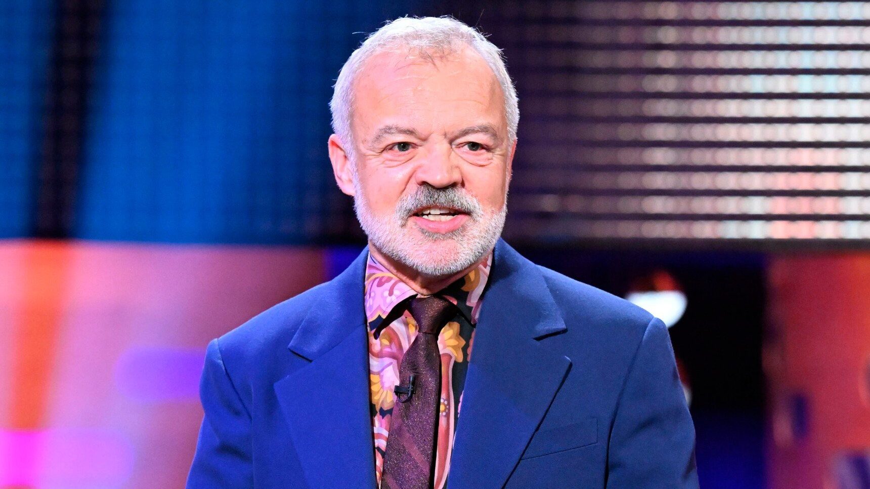 Talkshow host Graham Norton wearing a blue suit and patterned burgundy tie on set