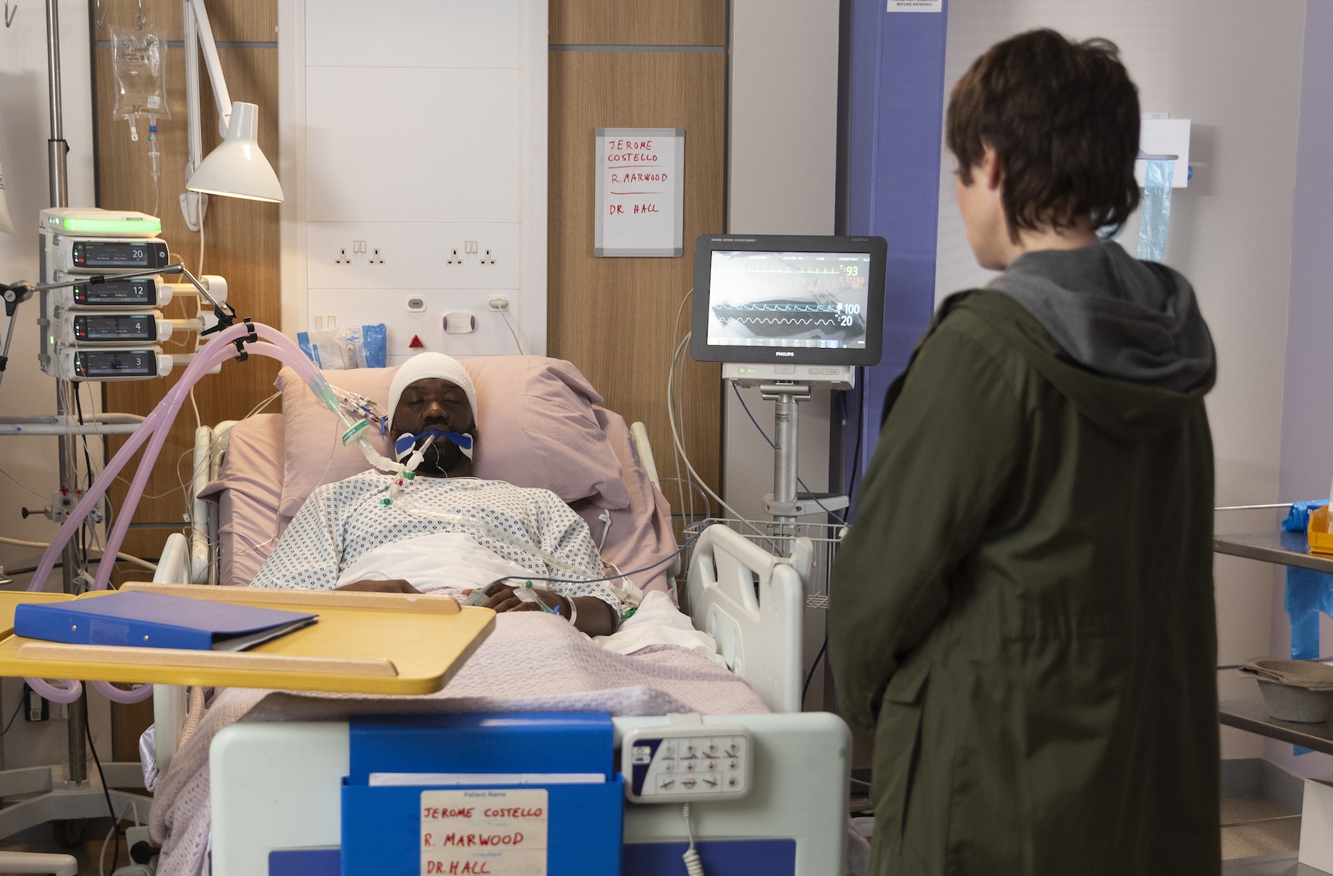 Costello in hospital bed with Becky watching him 