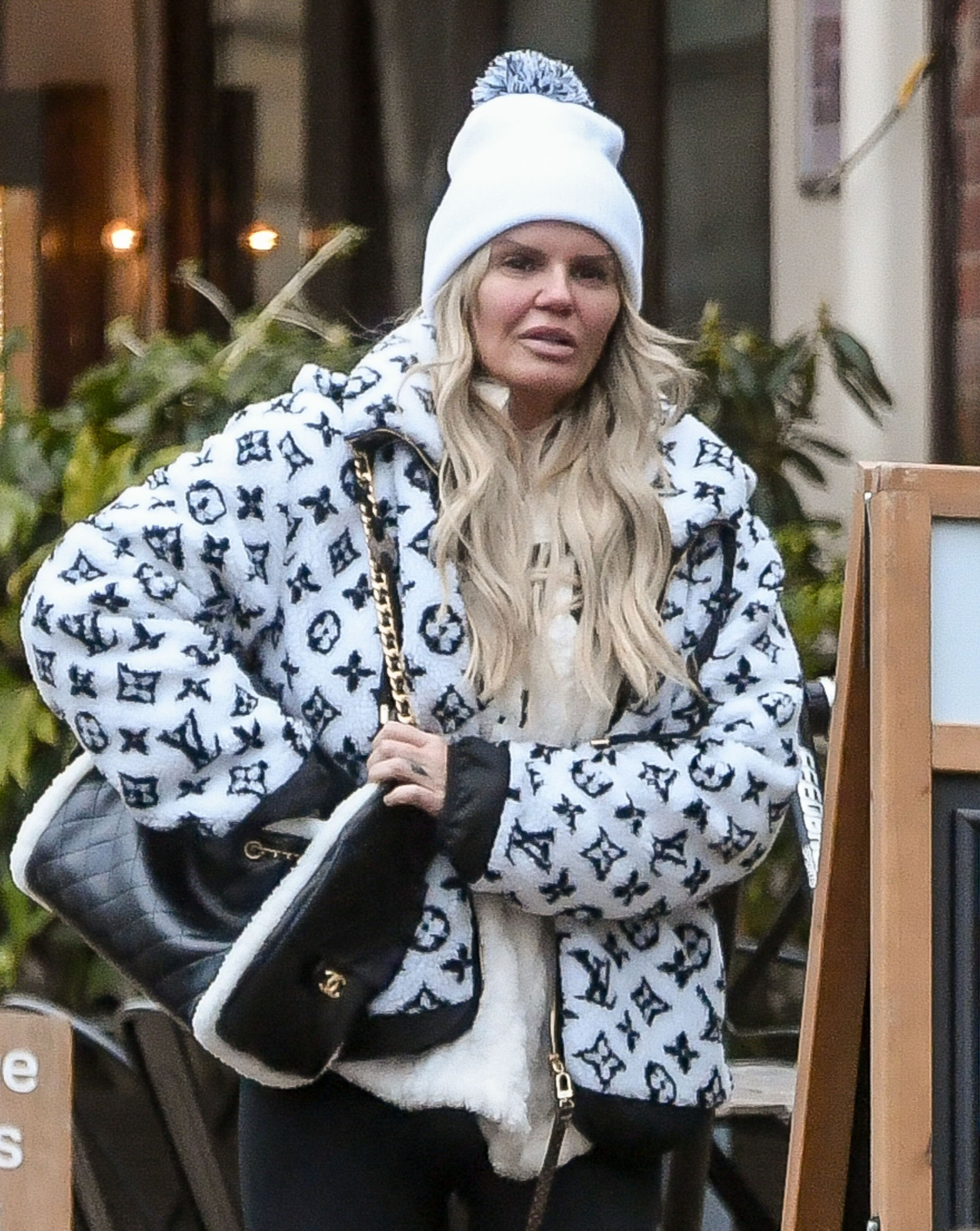 Kerry Katona wearing a patterned coat, bobble hat and handbag with serious expression