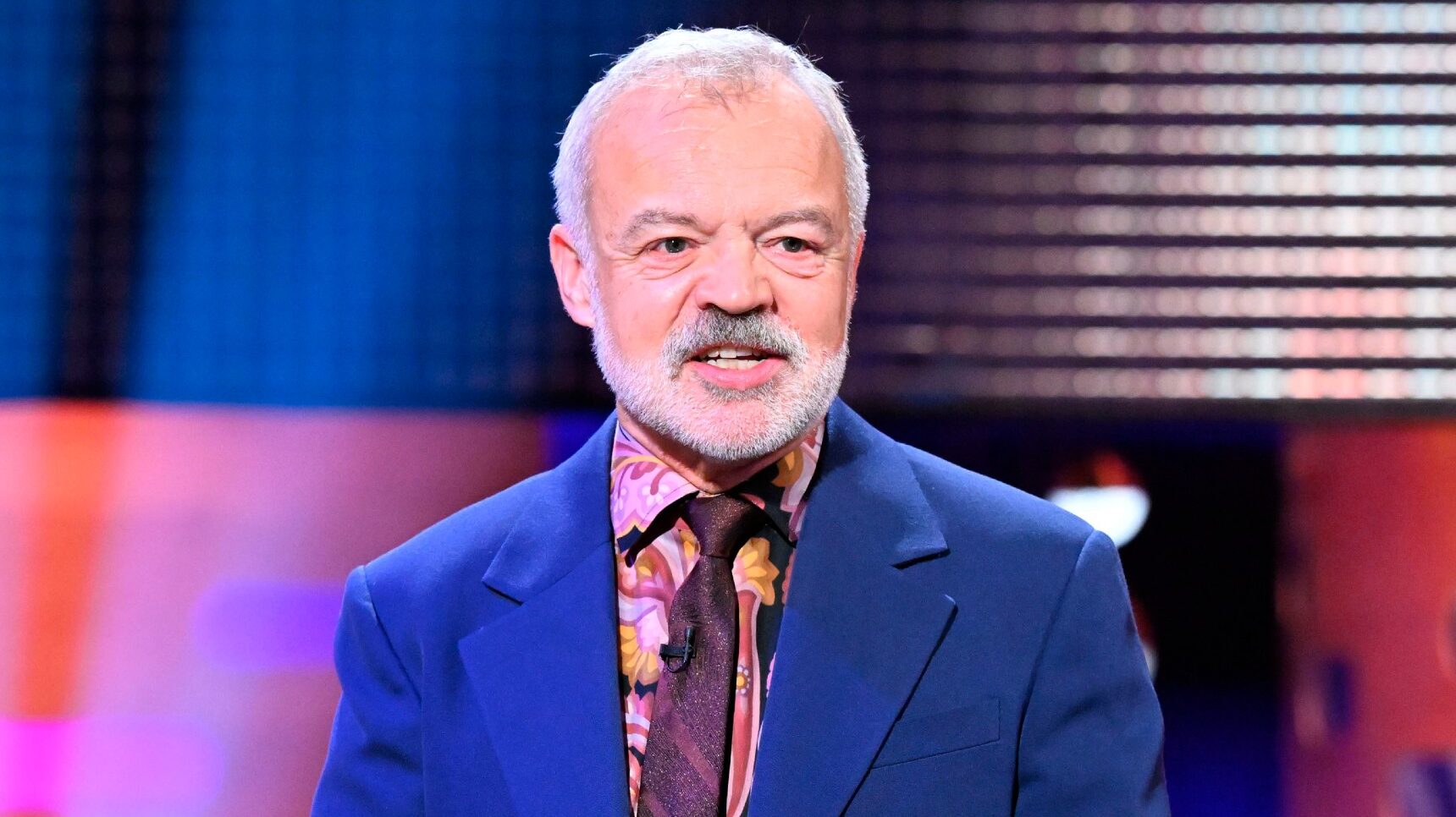 Talkshow host Graham Norton wearing a blue suit and patterned burgundy tie on set