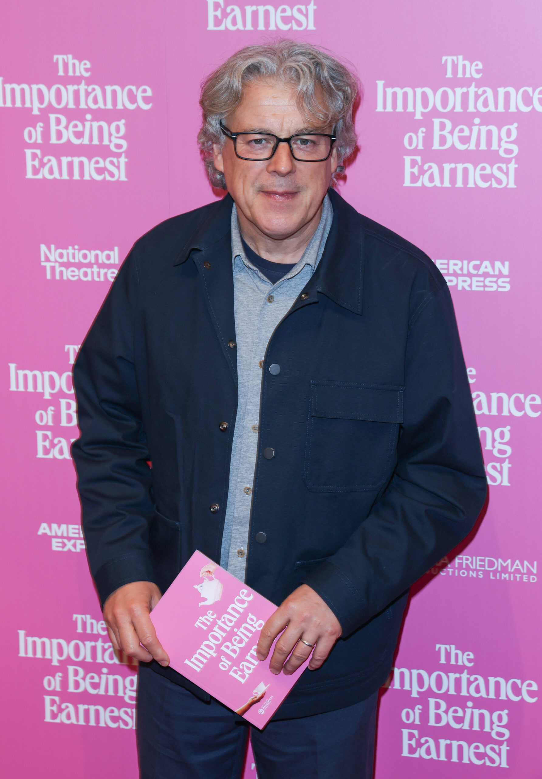 Alan Davies wearing glasses against a pink back board