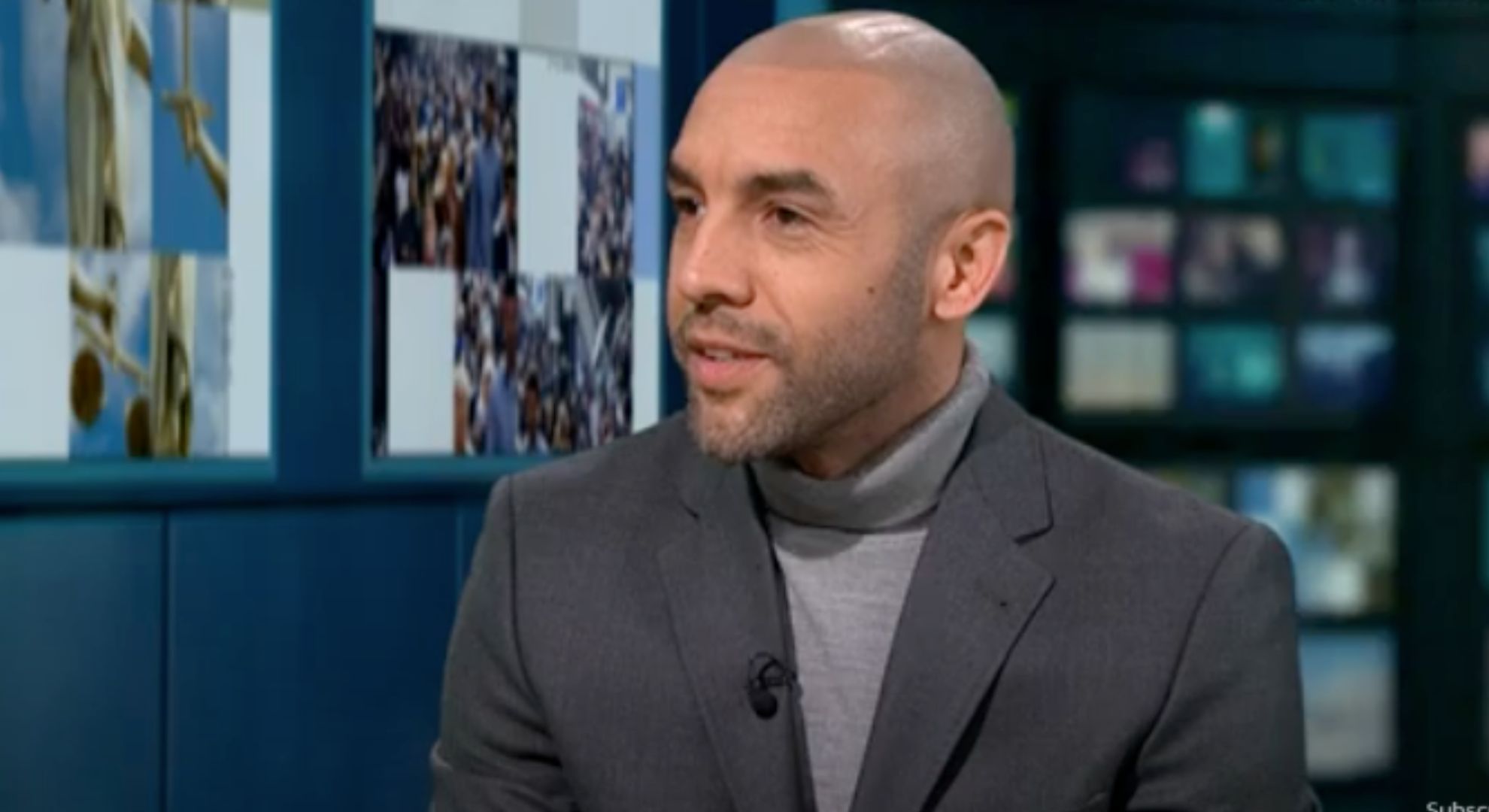 Alex Beresford smiles as he wears a dark grey suit and grey turtle neck jumper