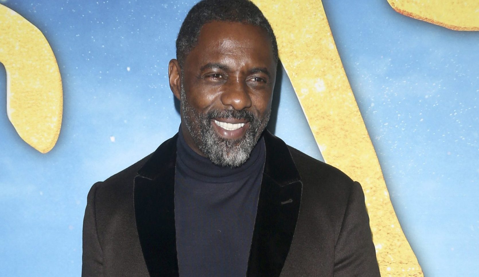 Idris Elba smiling wearing a dark jacket and polo neck
