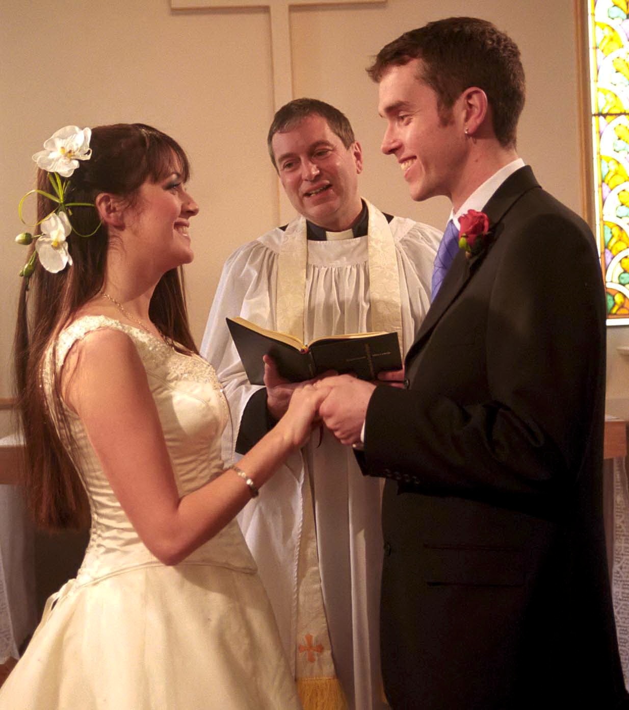 Tricia and Marlon smiling at each other as they get married with Ashley watching in Emmerdale
