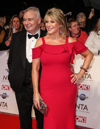 Ruth Langsford in a red gown poses with one hand on her hip looking to the side alongside husband Eamonn Holmes in a black tuxedo 