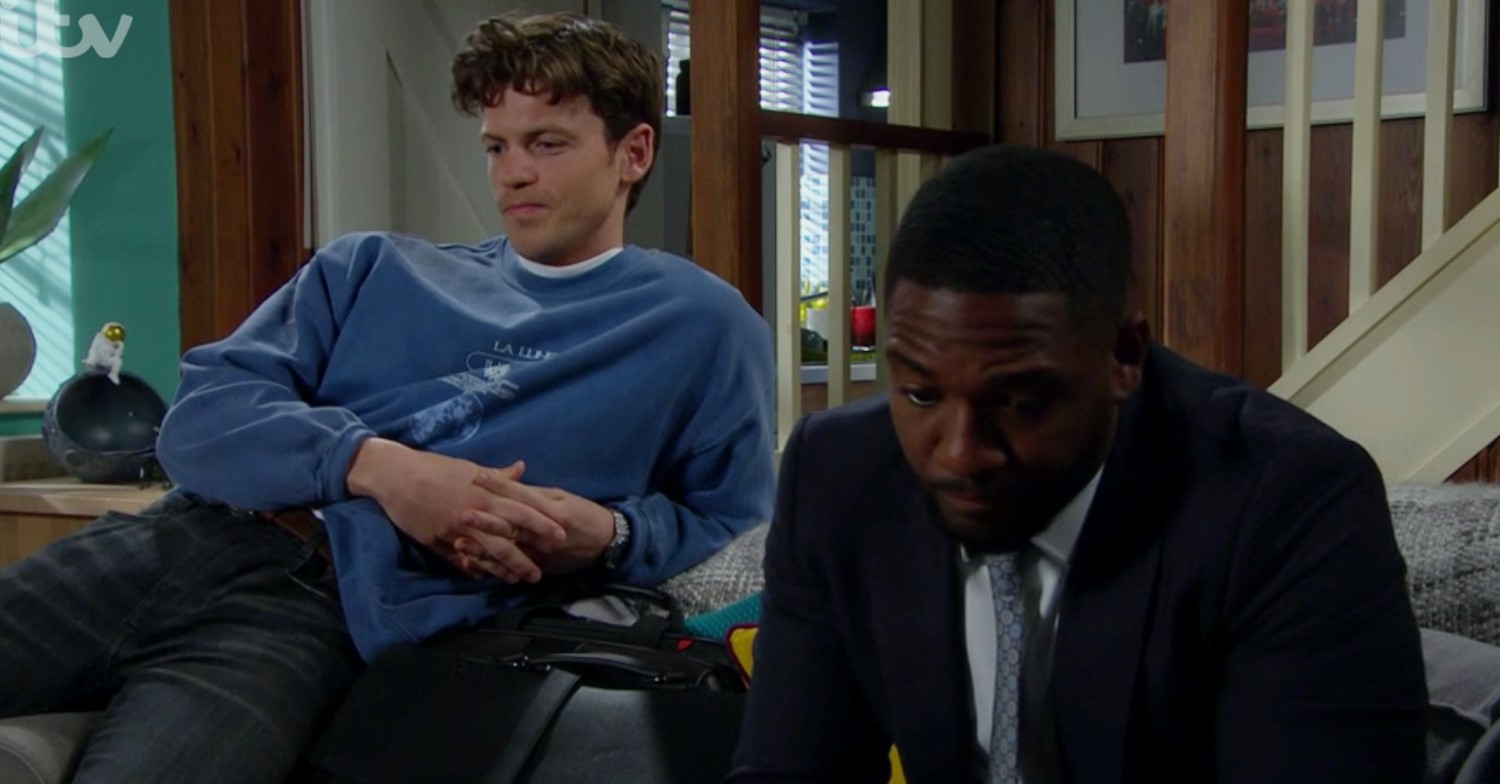Ethan Anderson and Marcus Dean sit on the sofa after an awkward conversation in Emmerdale