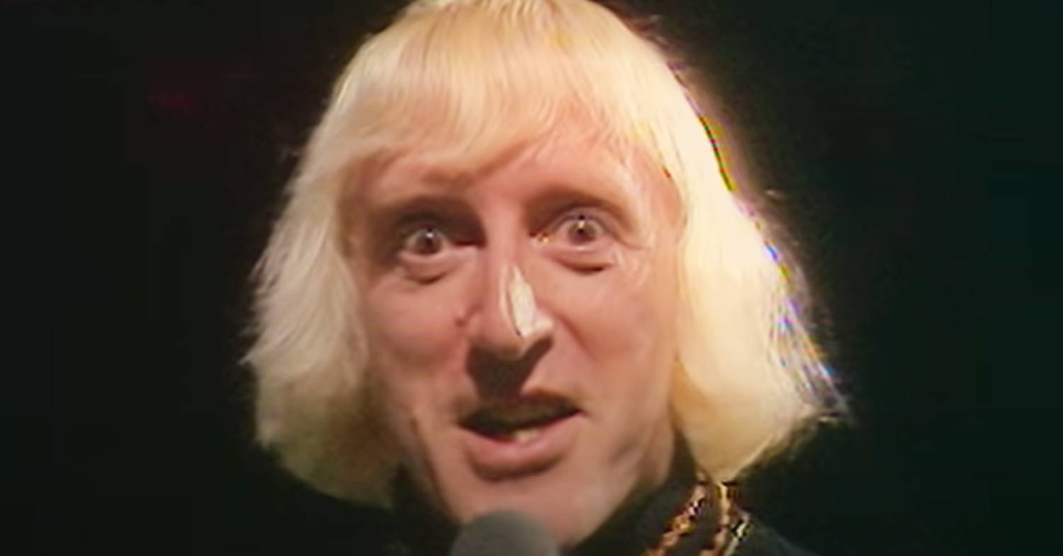 Jimmy Savile smirking at the camera on a dark background