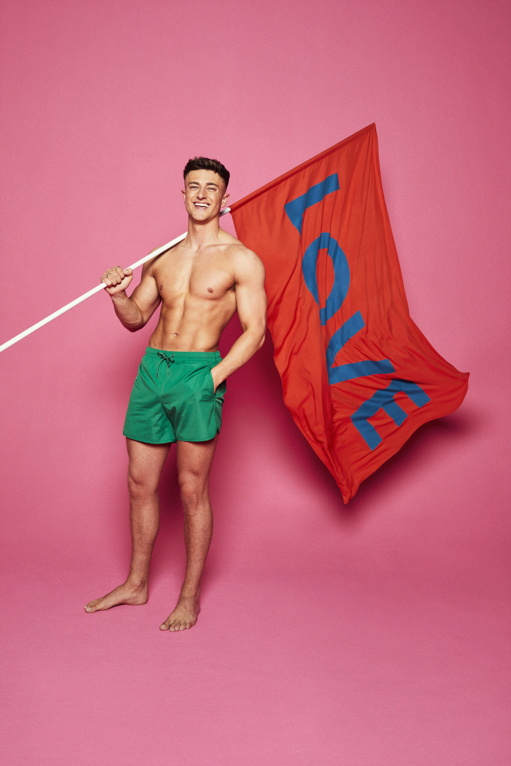 Love Island Liam smiling in green swim trunks while holding flag