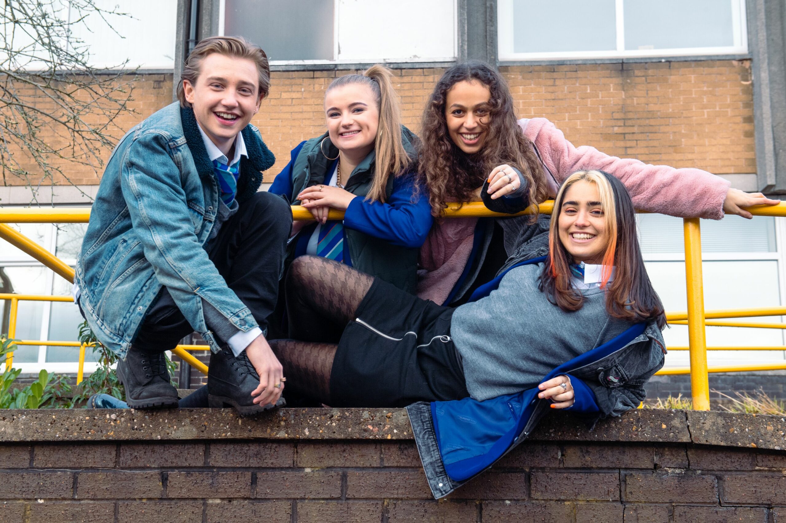 Ackley Bridge characters Johnny (Ryan Dean), Queenie (JASMINE PAYNE), Kayla Afzal (Robyn Cara), Fizza (Yasmin Al-Khudhairi) grinning on a school wall