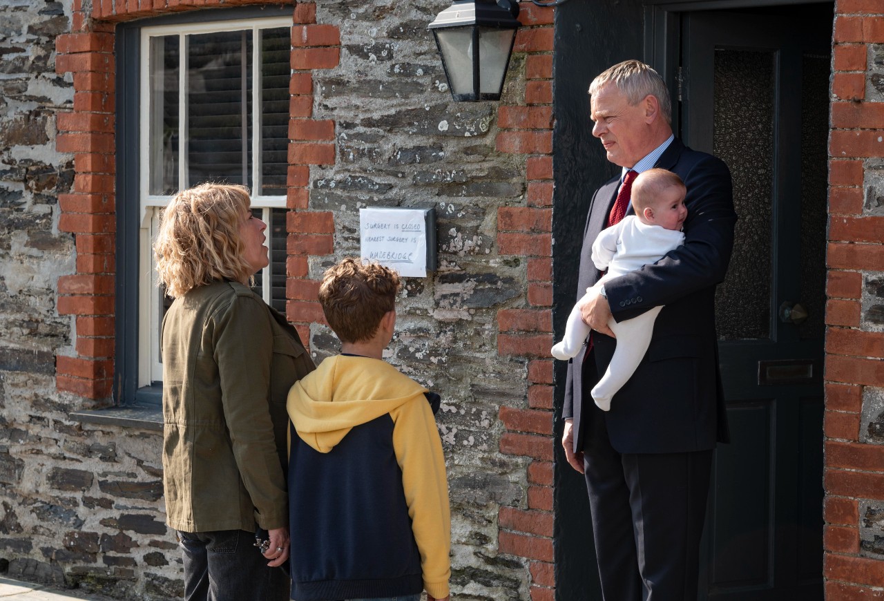 DOC MARTIN SEASON 10 EPISODE 1 PROMOTIONAL IMAGE Martin Clunes as Doc Martin holds a baby and looks down at a woman and child