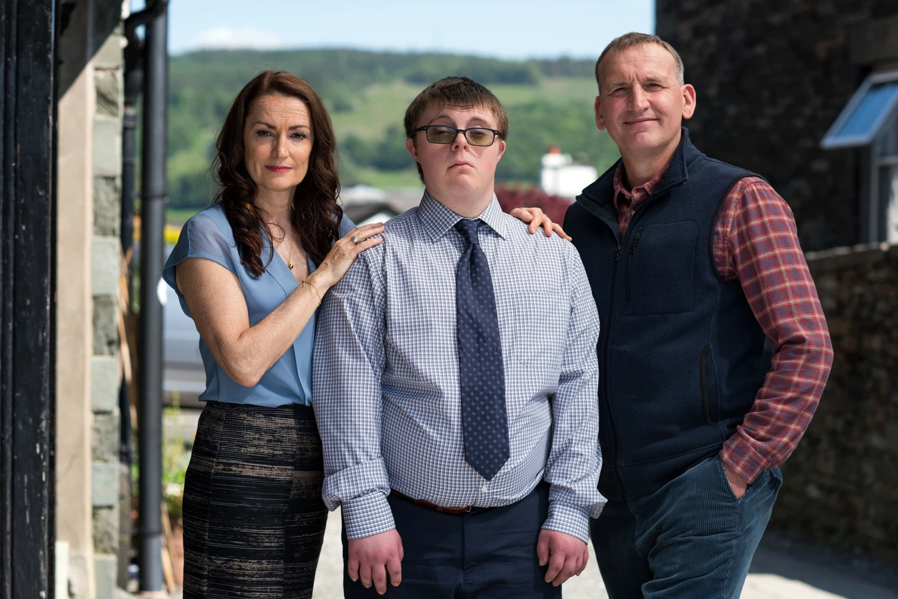 Pooky Quisnel as Louise (left), Leon Harrop (middle) and Christopher Eccleston as Maurice (right), they stand outside together smiling and hugging