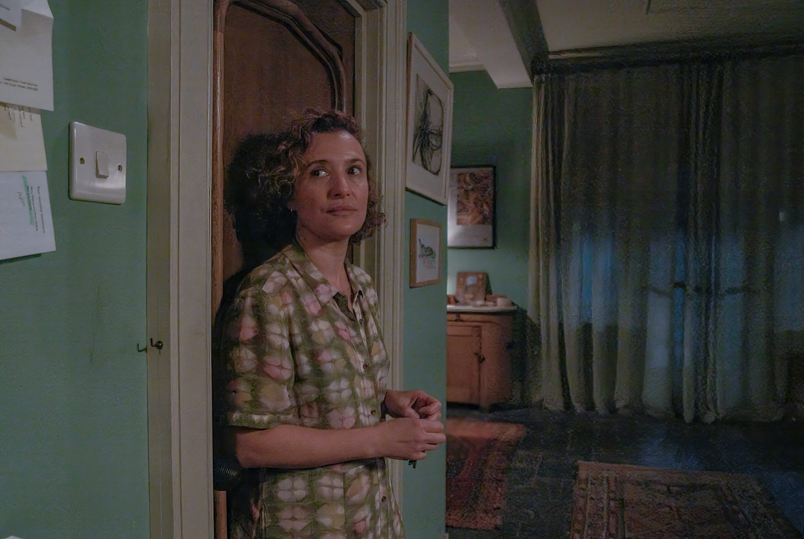 Lyndsey Marshall as Mary Watling, she leans against a door in her home, she has a plaid shirt on and a small smile