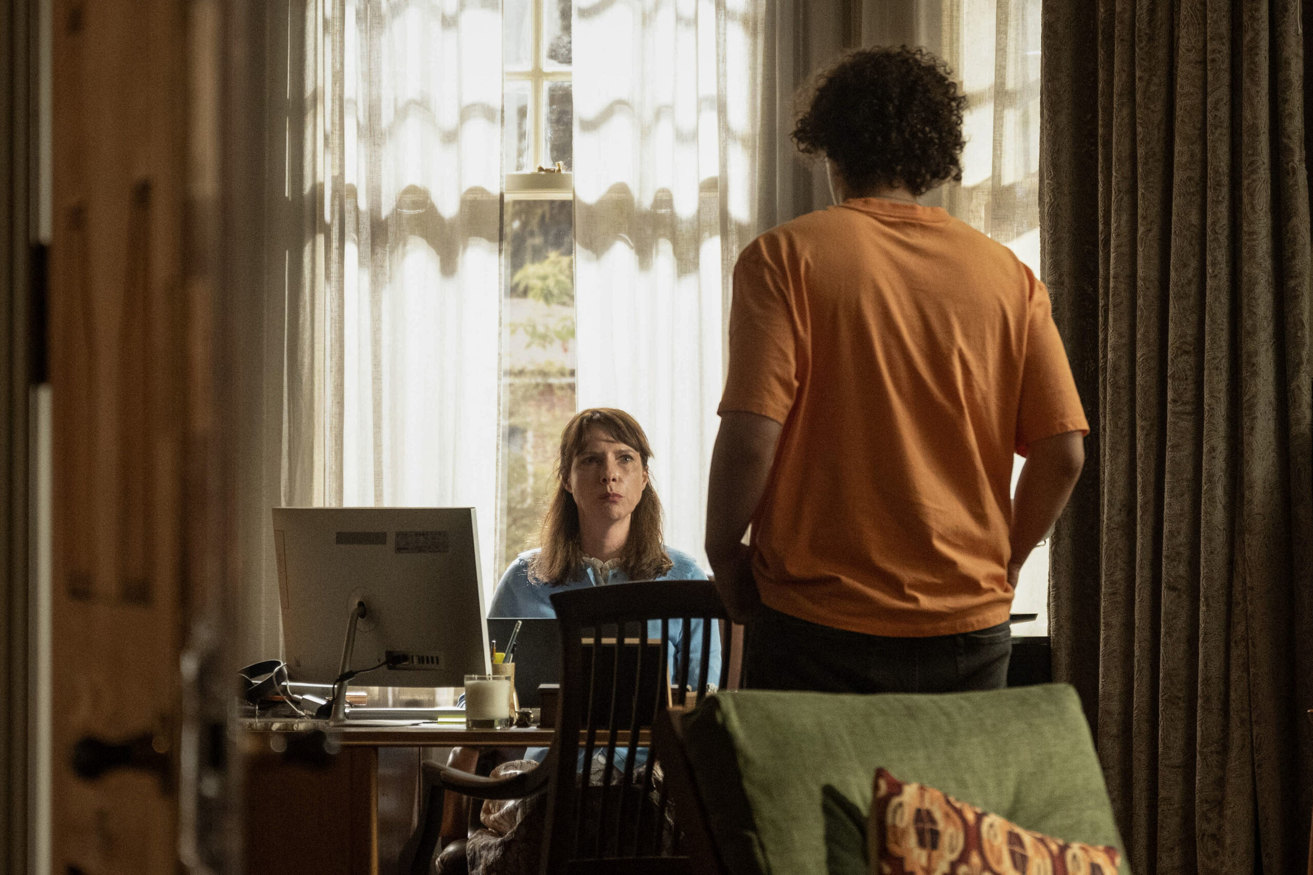 Dolly Wells as Janice Fife, she sits at a desk with a computer as Ben Watling stands infront of it