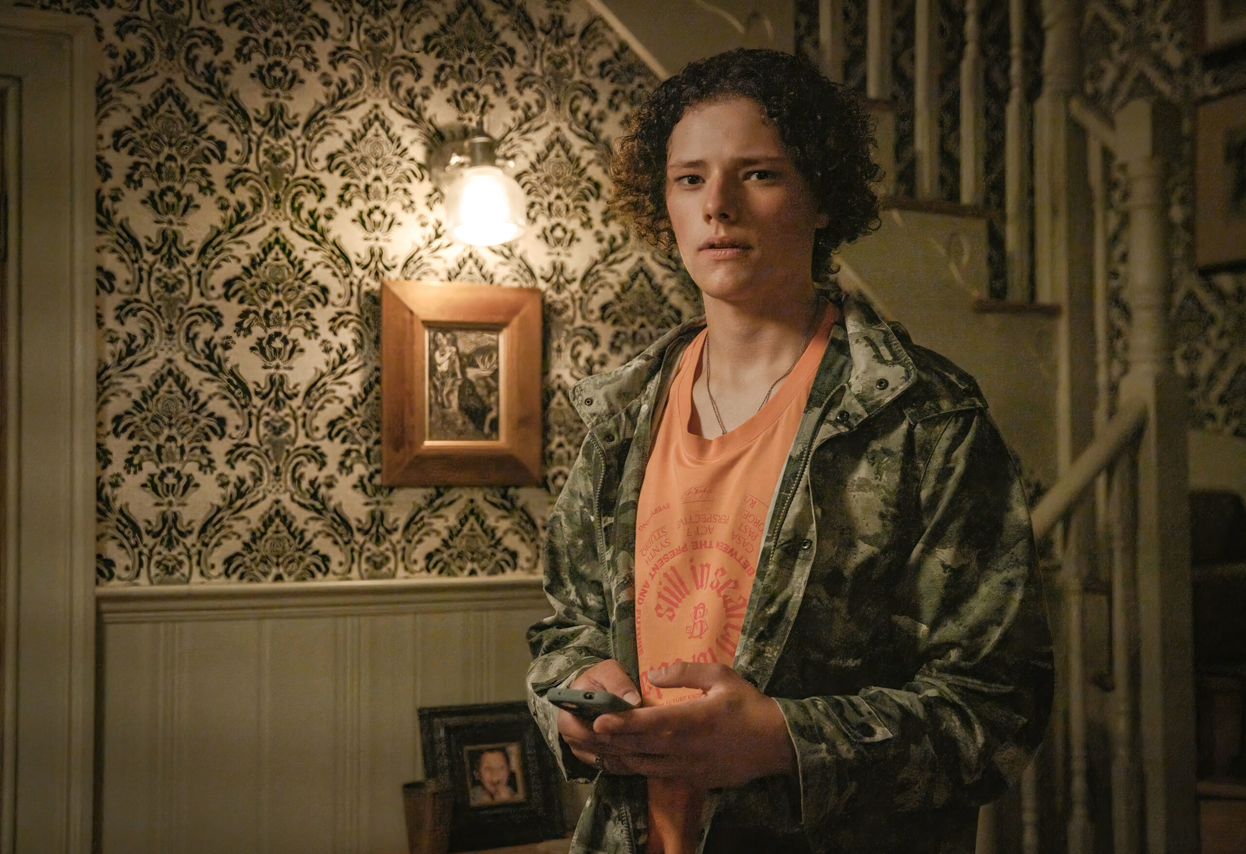 Louis Oliver as Ben Watling, he stands in his hallway with his phone in his hands in Inside Man