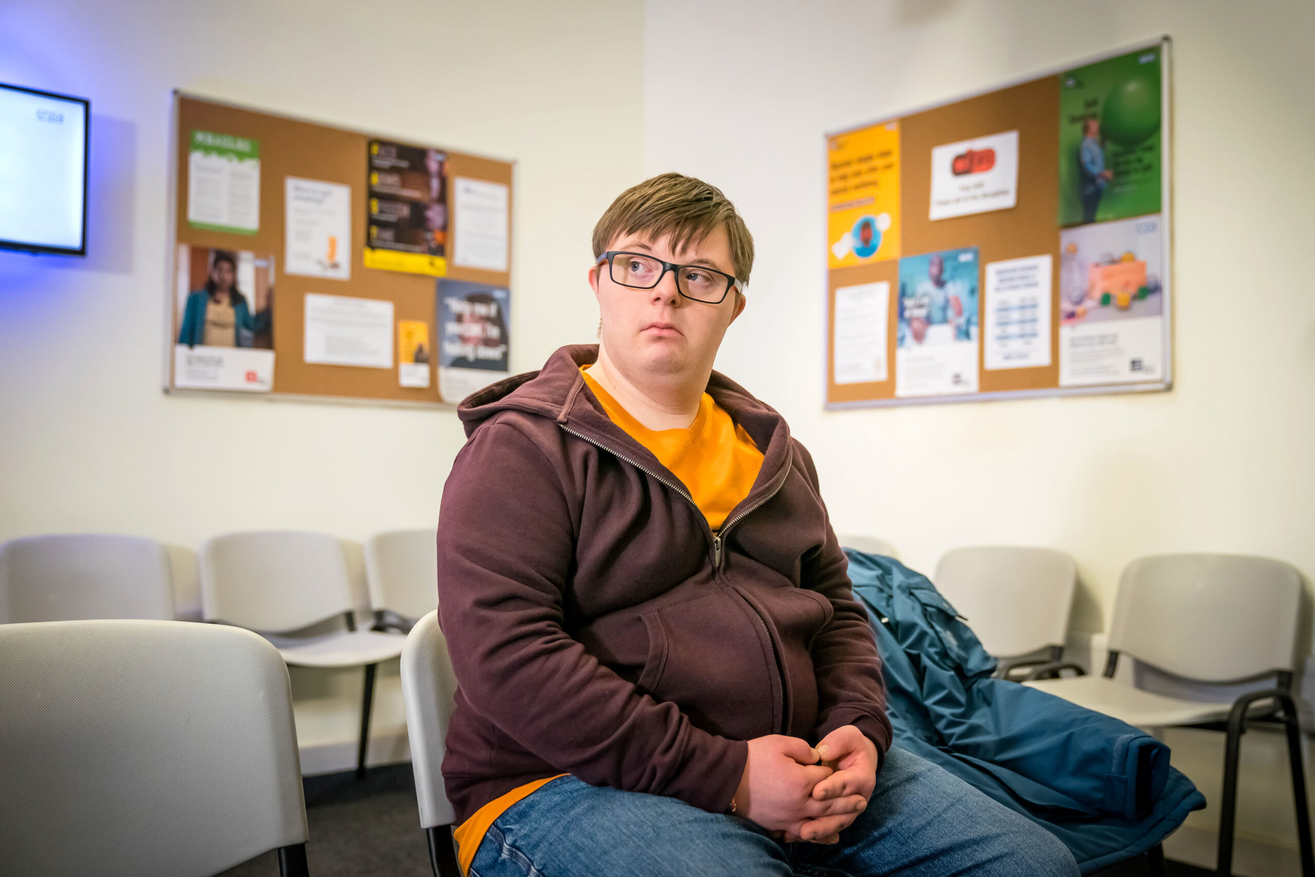Leon Harrop as Ralph Wilson in Ralph & Katie, he sits in a room with chairs with a sad face with his hands together