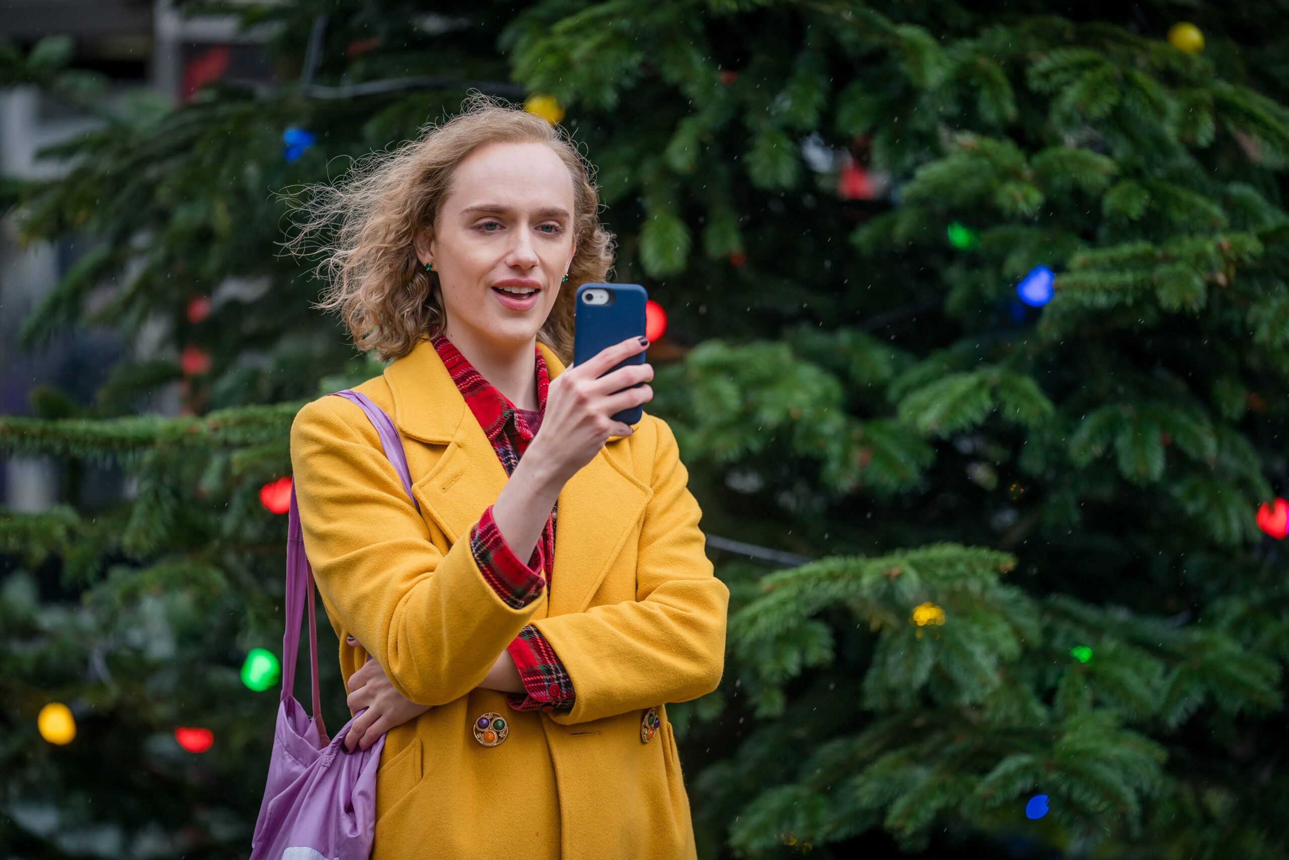 Matt Greenwood as Tom, he stands next to a christmas tree in a yellow coat with his phone in front of him, he is smiling
