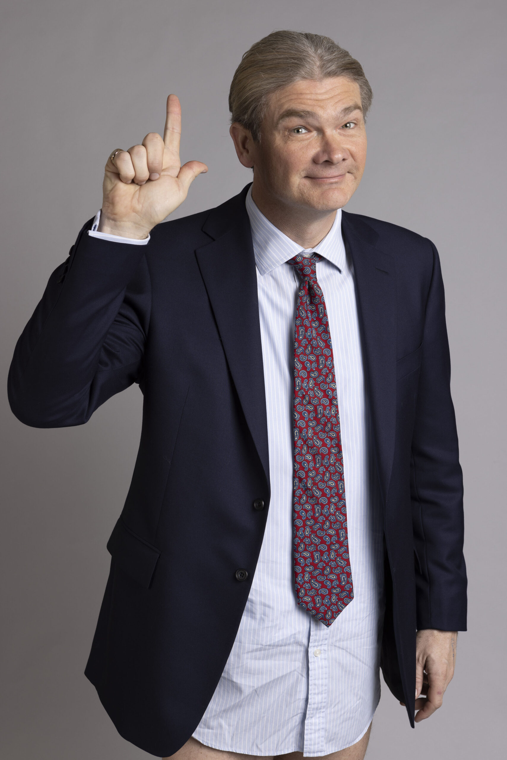 Simon Farnaby as Julian, he is in a shirt and tie, he points up with a smile