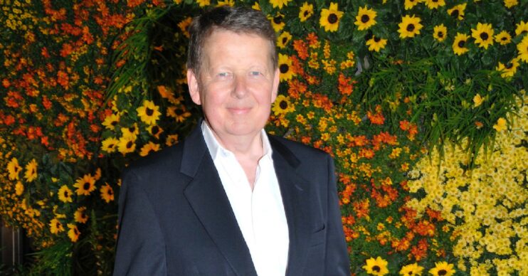 Bill Turnbull in a dark suit in front of a wall of flowers