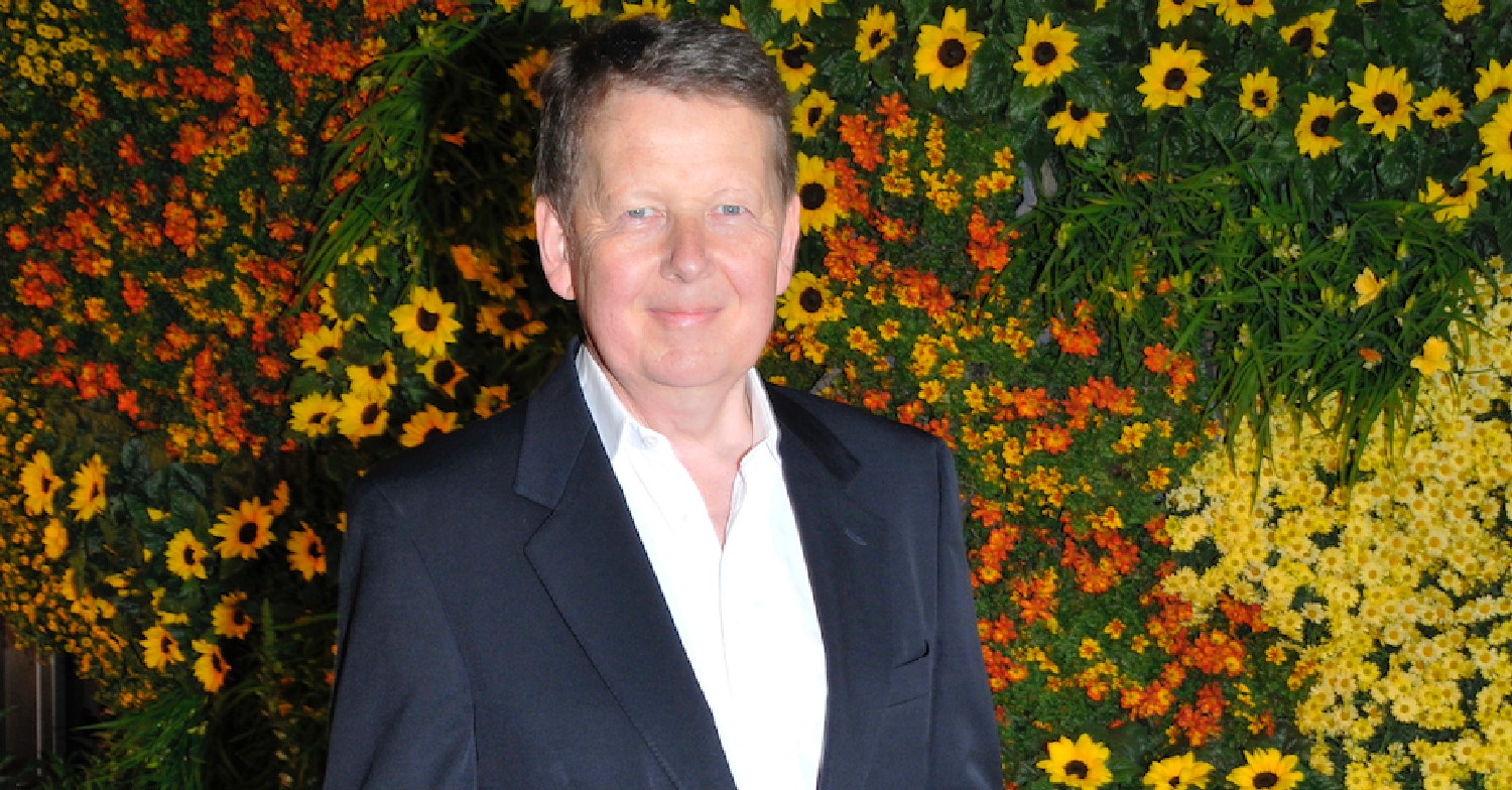 Bill Turnbull in a dark suit in front of a wall of flowers