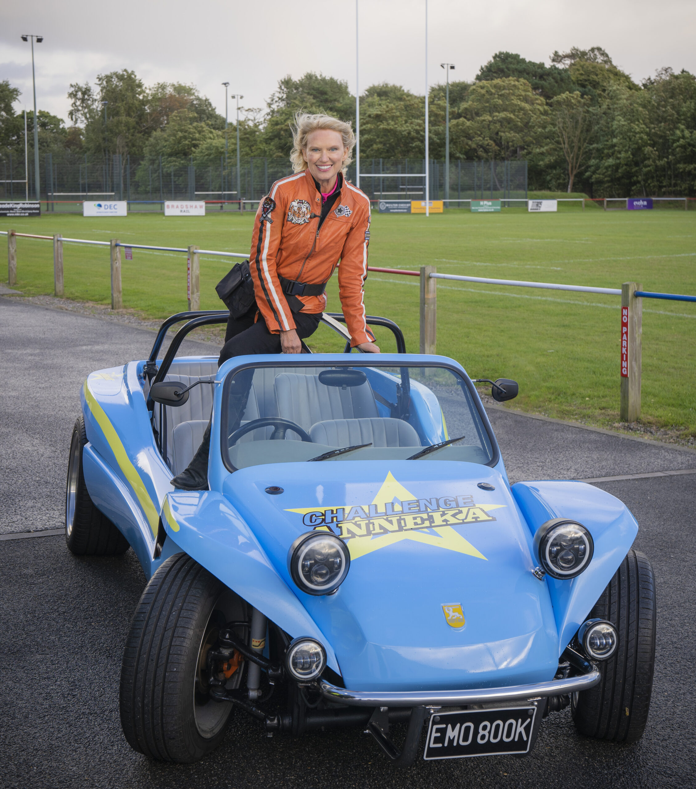 Anneka Rice in Challenge Anneka, she stands outside in an orange coat on top of a buggy
