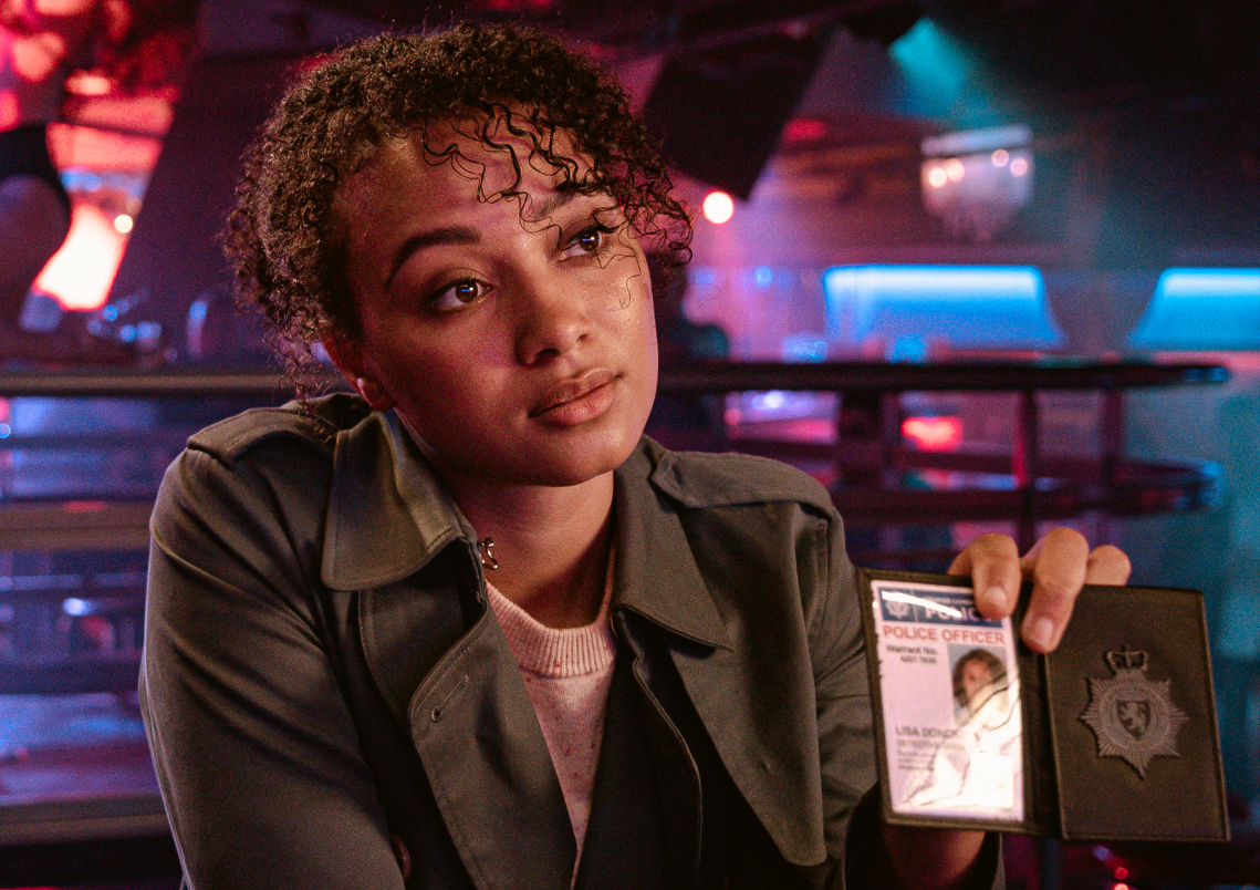 Emma Naomi as Lisa Donkers, she holds up her policing ID in a venue, she has a green coat on