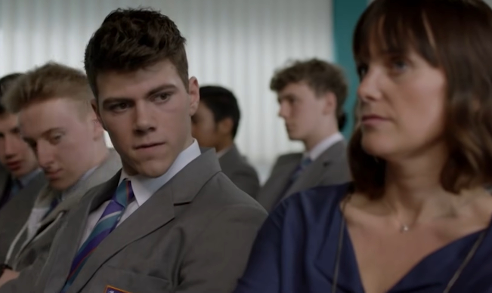 Sam Retford as Cory Wilson from Ackley Bridge, he sits in an assembly next to a teacher shooting a disapproving look to his right