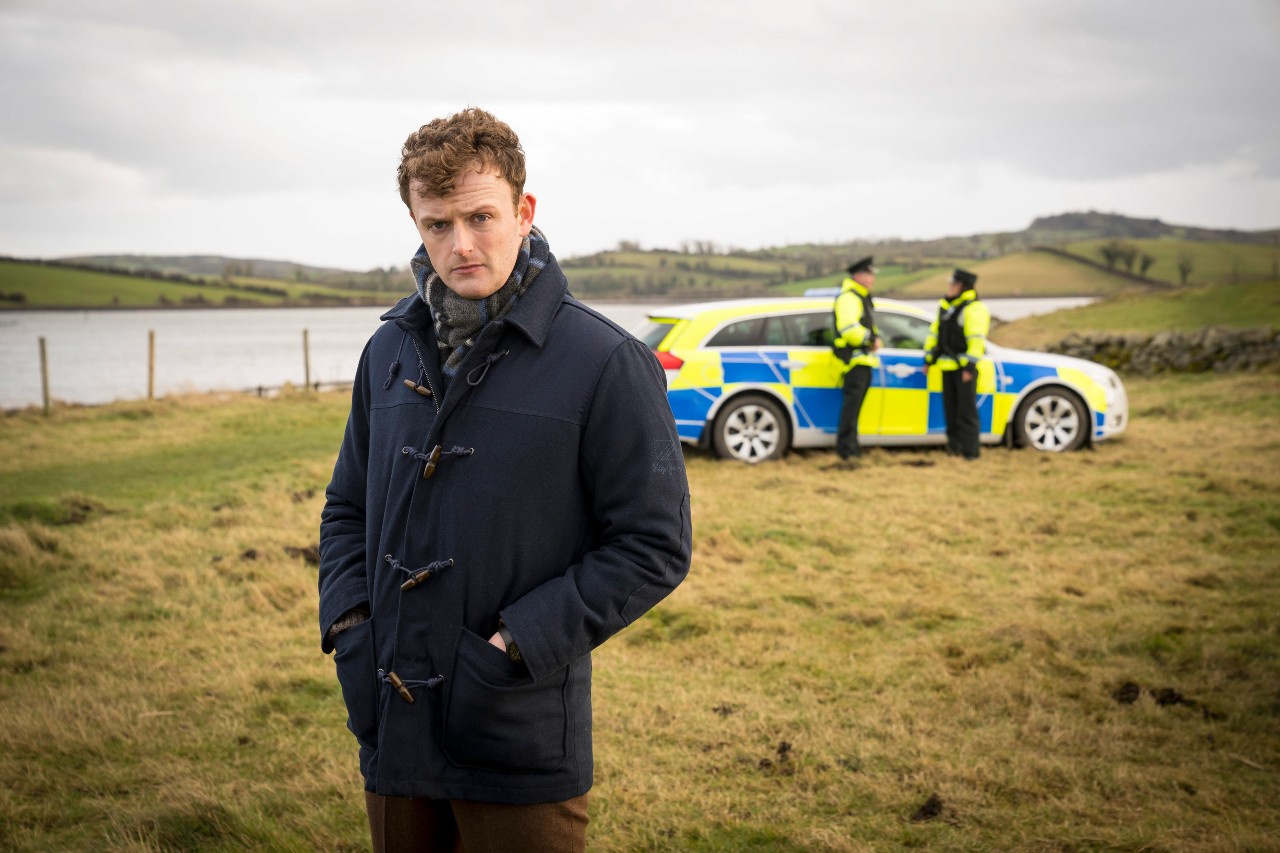 CHRIS WALLEY AS DS BIRDY, he stands in a field with a PSNI car behind him