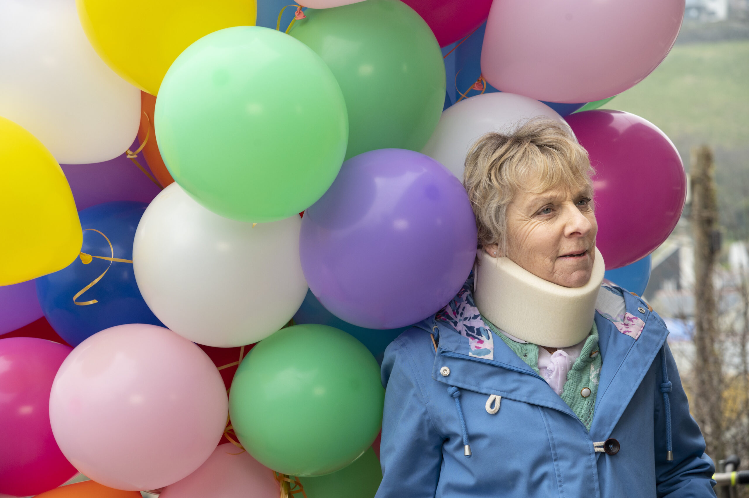 Mrs Tishell wearing a neck brace and surrounded by balloons in Doc Martin